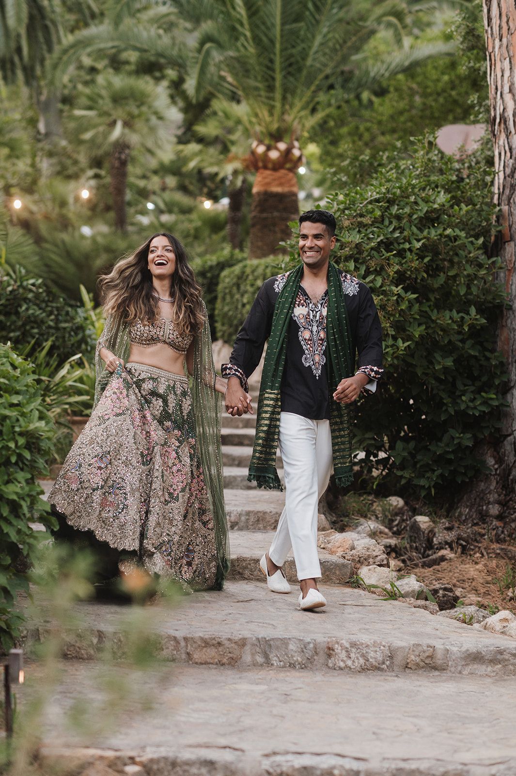Couple in traditional Indian wedding attire walk hand-in-hand through a lush garden, laughing joyfully together.