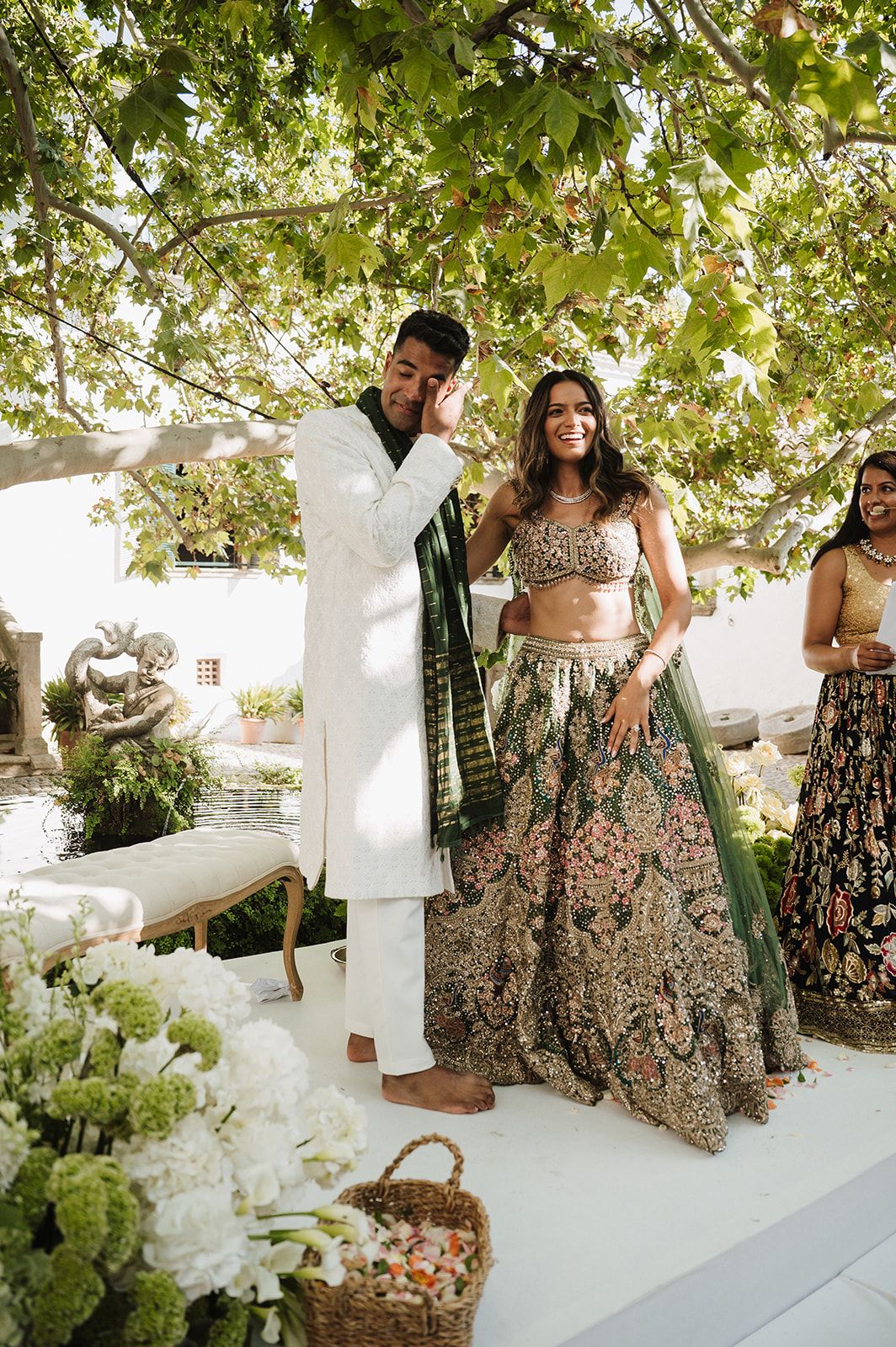 Bride in green floral lehenga stands with groom in white sherwani during outdoor Indian wedding ceremony.