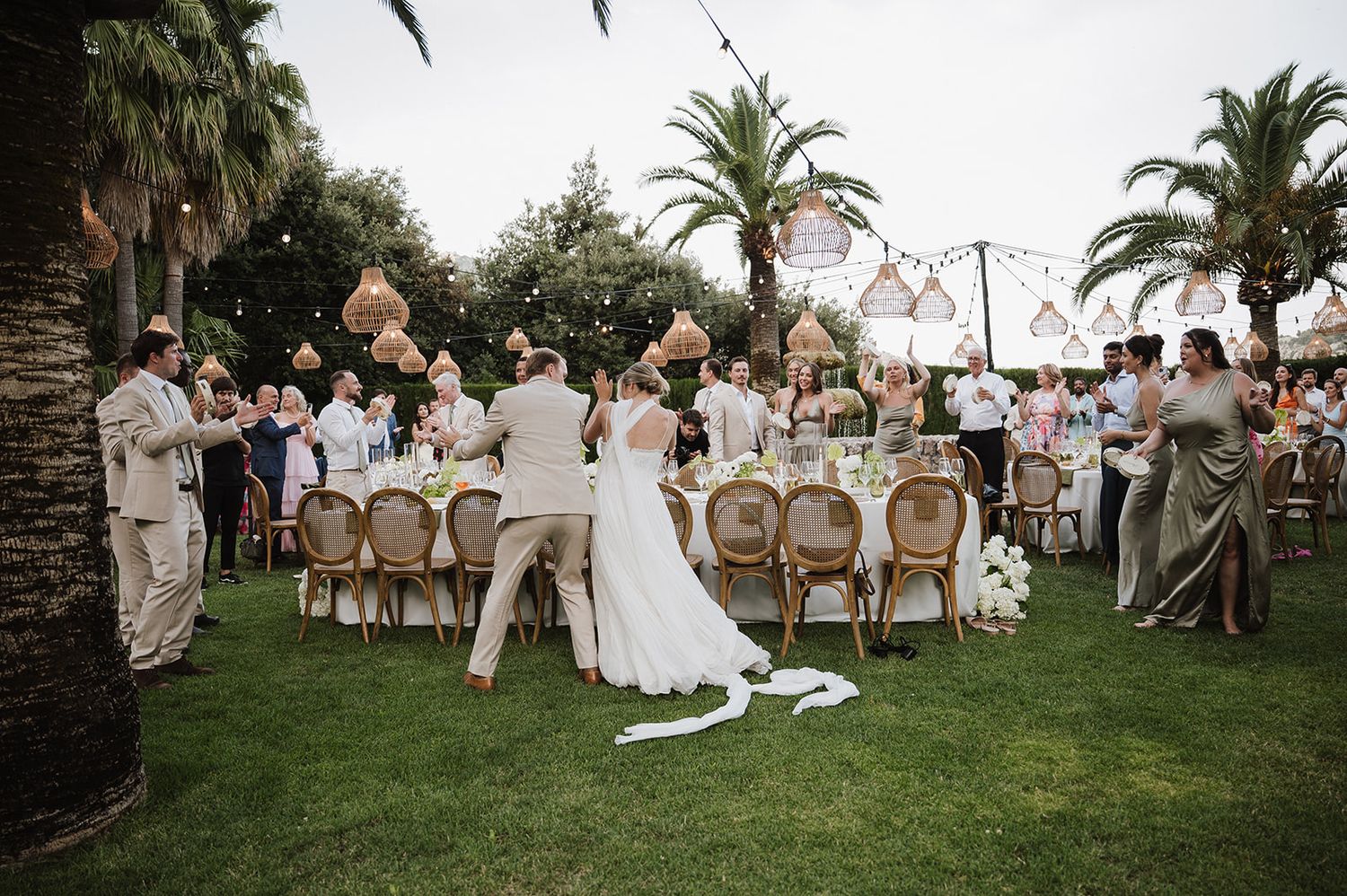 Bride and groom celebrate with guests at an outdoor evening reception surrounded by palm trees and string lights.