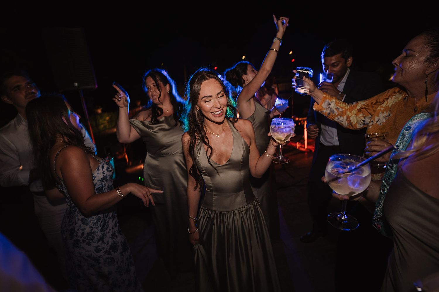 Woman in sage green dress dancing with arms raised at lively outdoor wedding reception party at night.