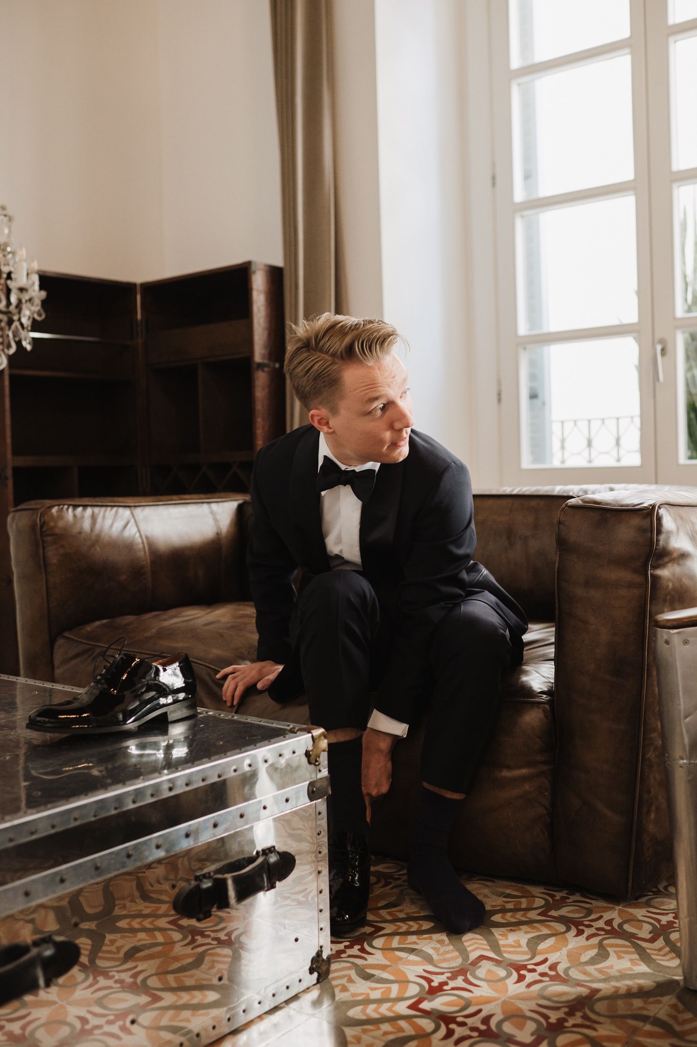 Groom in black tuxedo sitting on leather sofa preparing for wedding ceremony.