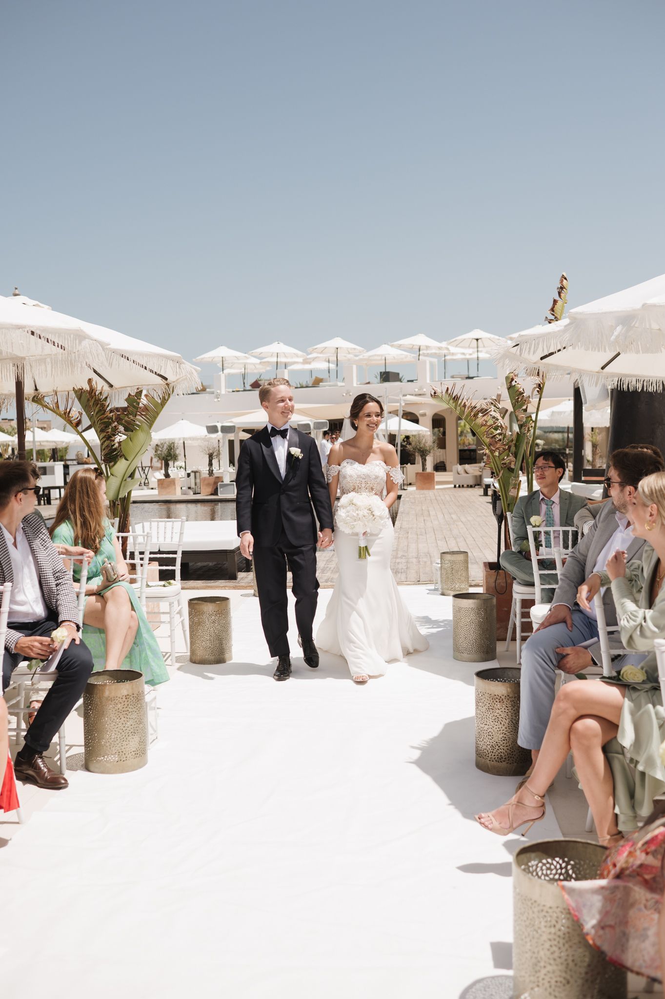 Bride and groom walk down outdoor aisle at sunny beach wedding ceremony with white umbrellas.