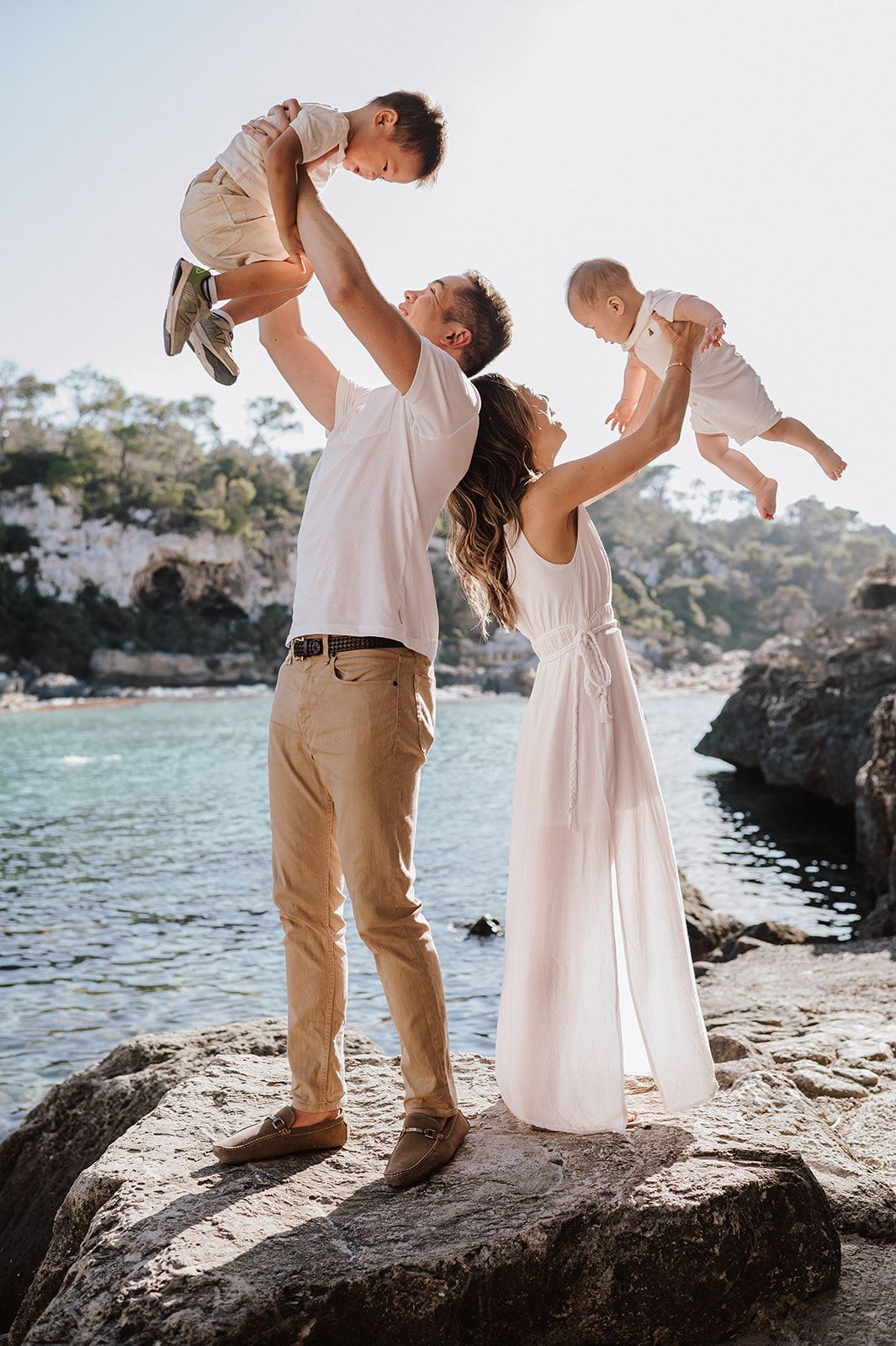 Couple lifting two babies in the air on rocky coastal shore with turquoise water backdrop.