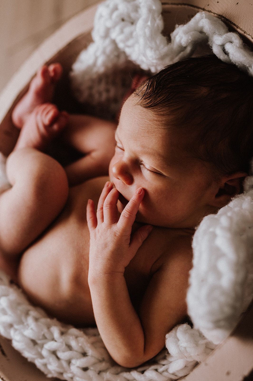 Peaceful newborn baby sleeping curled up in a wooden bowl with white chunky knit blanket and bunny ear props.