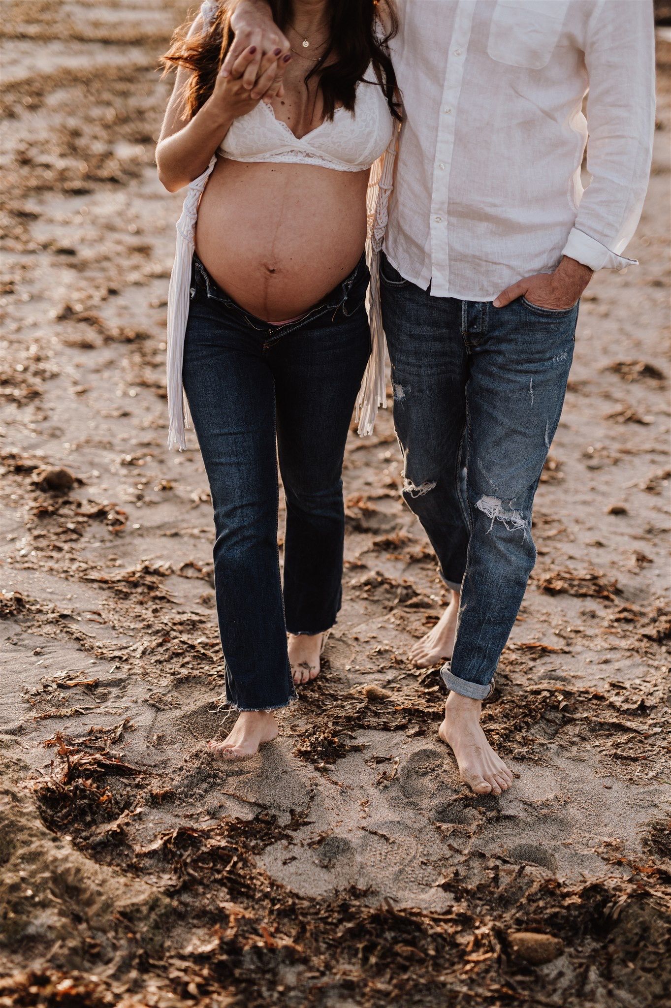 Pregnant woman in white bralette and jeans standing barefoot on beach sand with her partner.