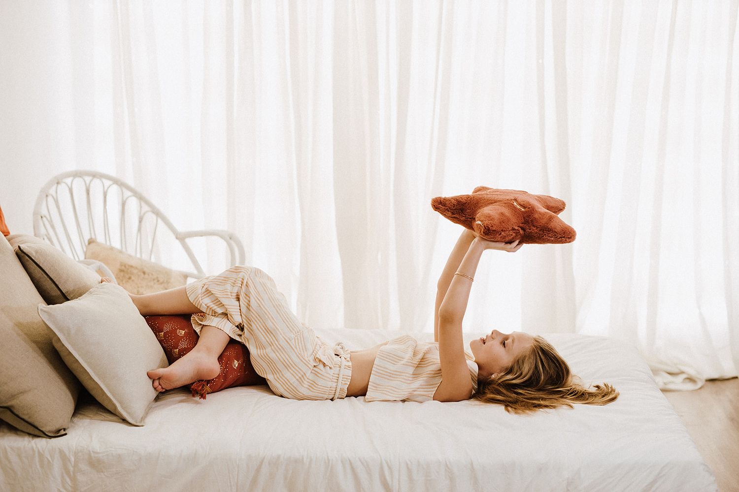 Young girl lying on a white bed playfully holding a brown stuffed animal up in the air.