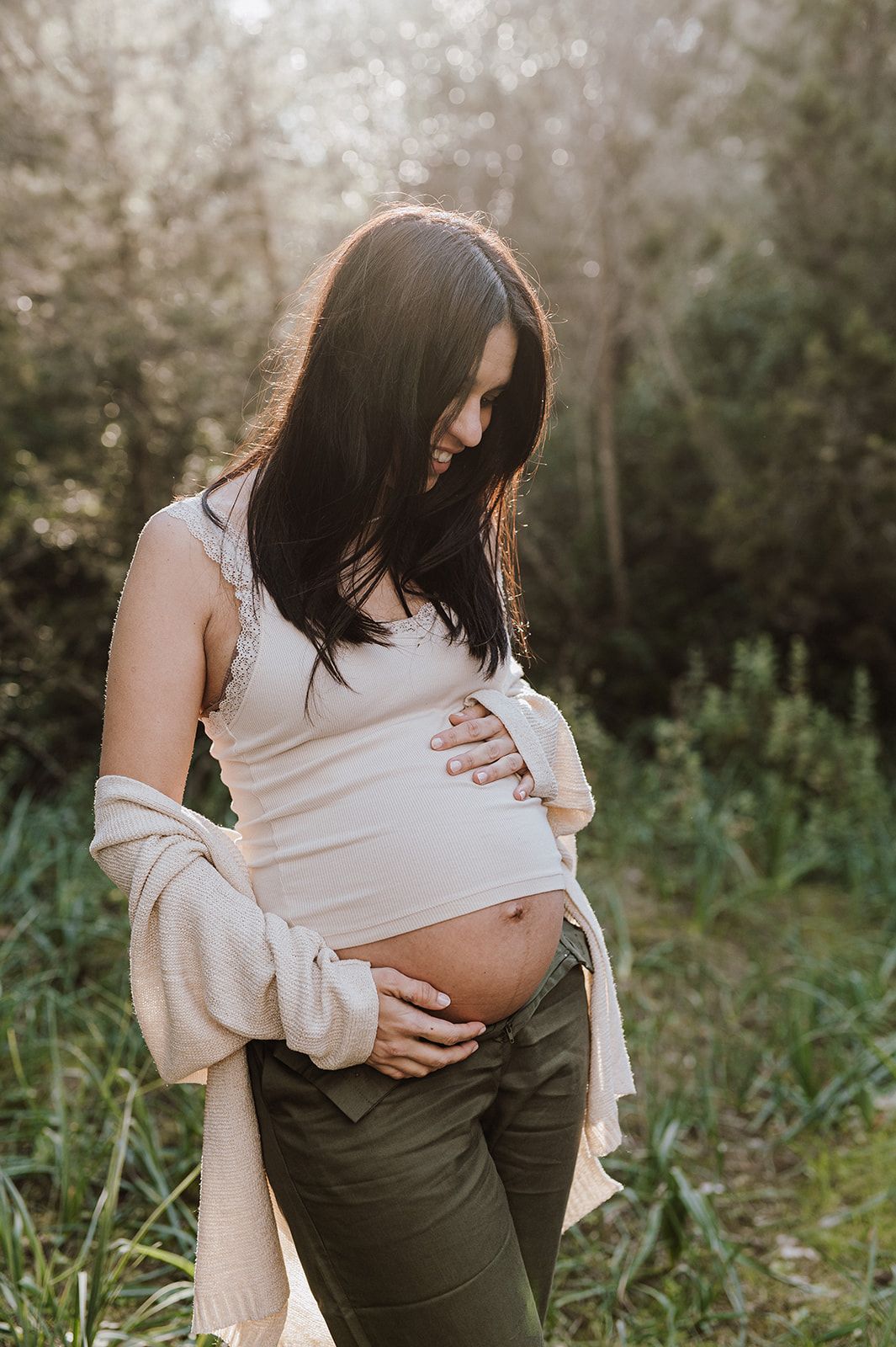 Smiling pregnant woman in white crop top and green pants cradling her baby bump outdoors in nature.
