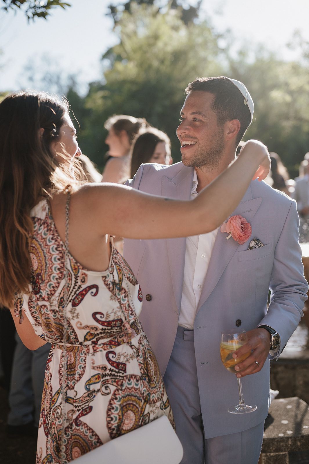 Groom in light gray suit with pink boutonniere laughing with guest at sunny outdoor wedding reception.