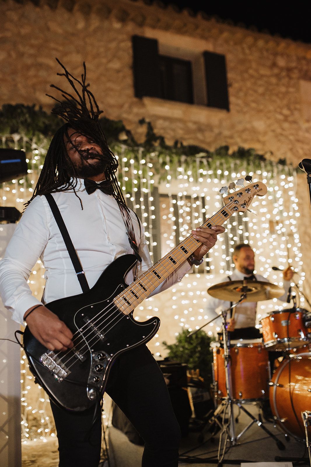 Energetic guitarist with dreadlocks performs passionately at an outdoor wedding reception with fairy light backdrop.