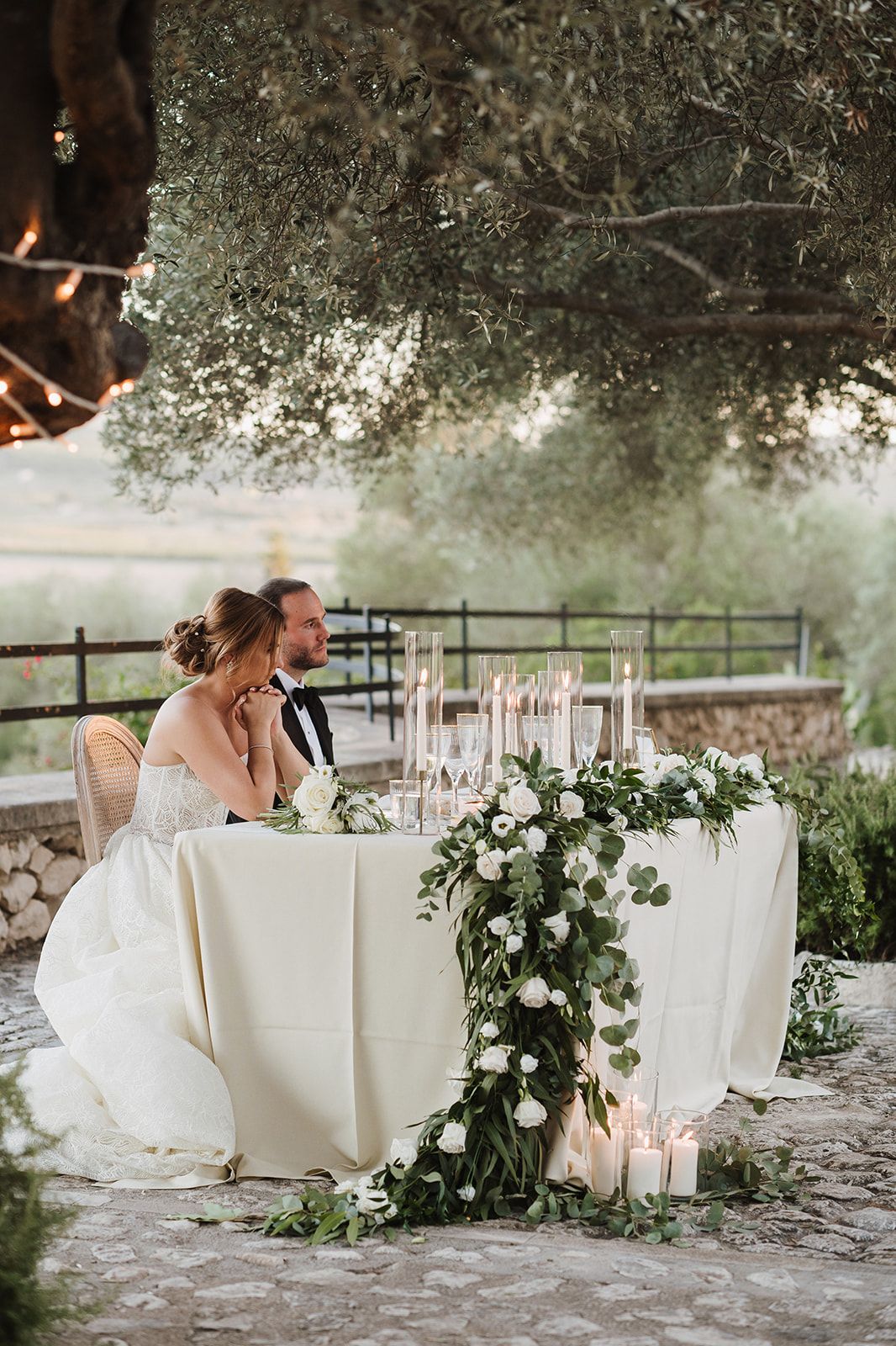 Bride and groom seated at sweetheart table adorned with white roses and candles beneath an olive tree.
