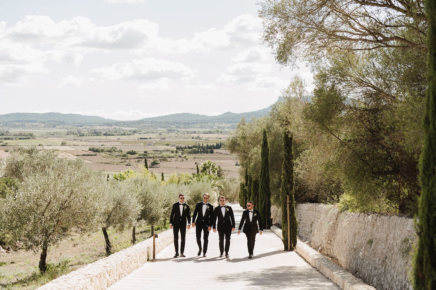Four groomsmen in black suits walk down a scenic road with rolling hills and cypress trees in background.