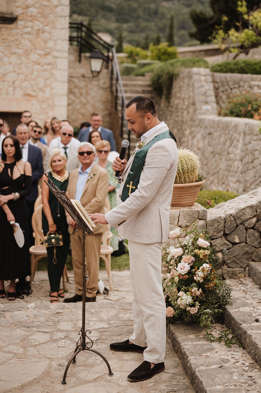 Wedding officiant in green stole reads from book during outdoor ceremony at a rustic stone venue.