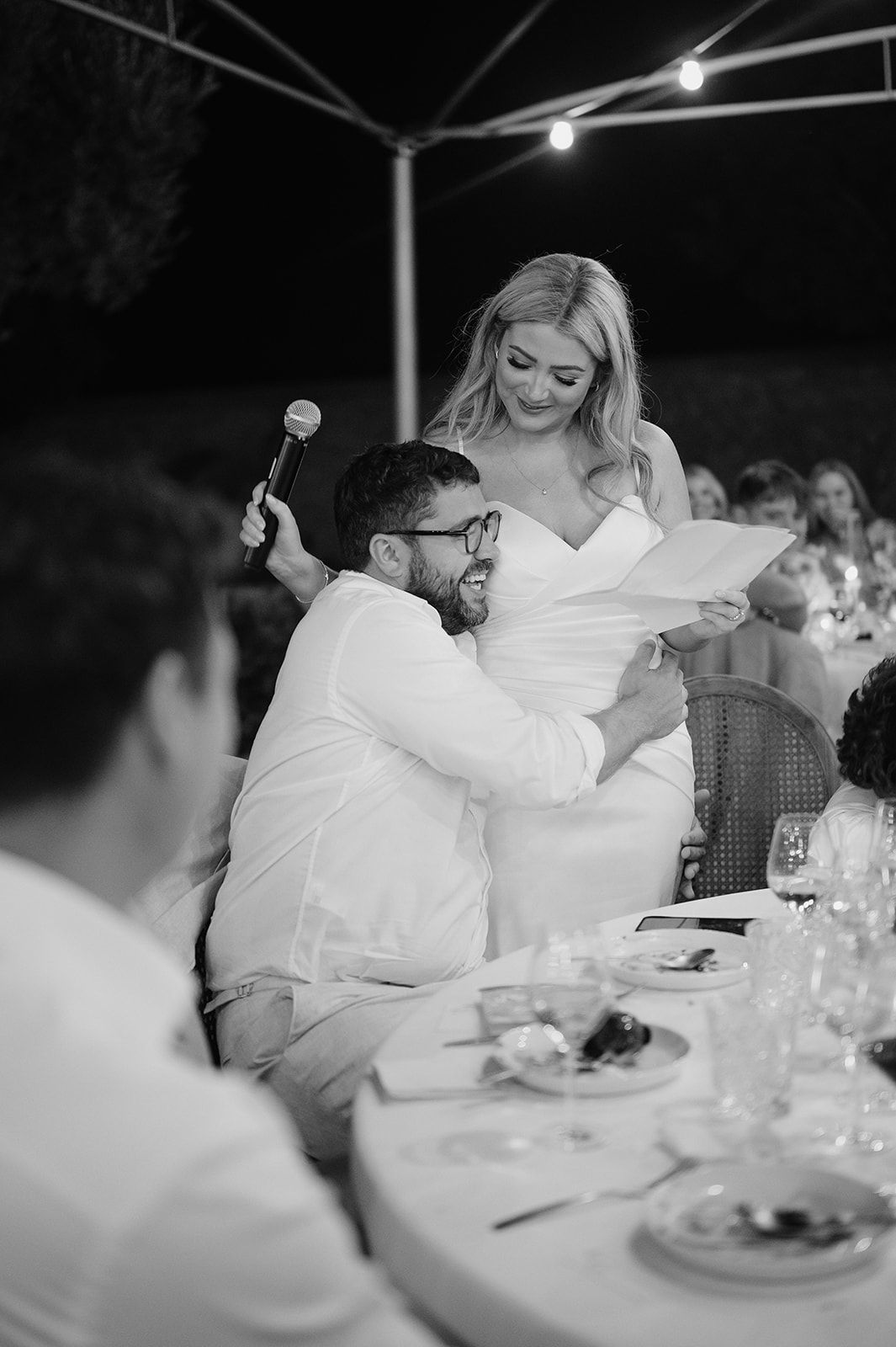 Bride reading speech while groom laughs and holds microphone at outdoor evening wedding reception.