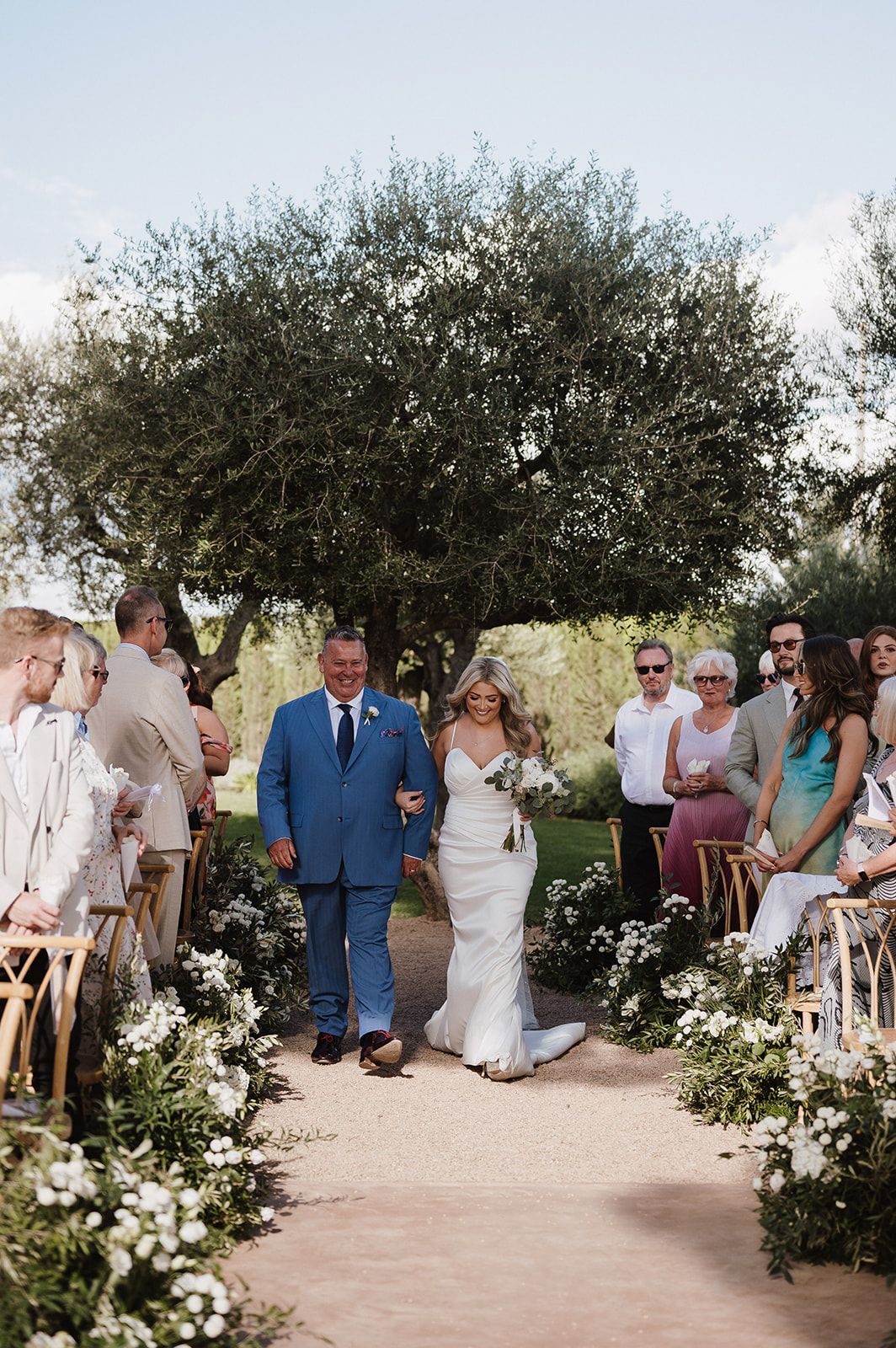 Bride walks down outdoor aisle with escort, flanked by white floral arrangements and seated guests.