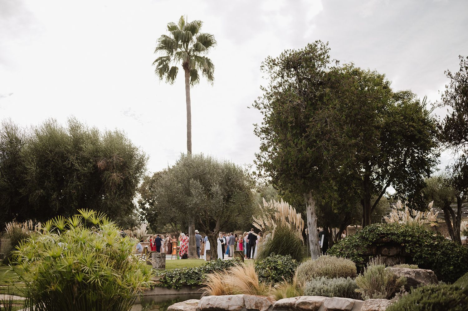 Wedding guests gather in a lush garden with palm trees and Mediterranean landscaping for an outdoor ceremony.