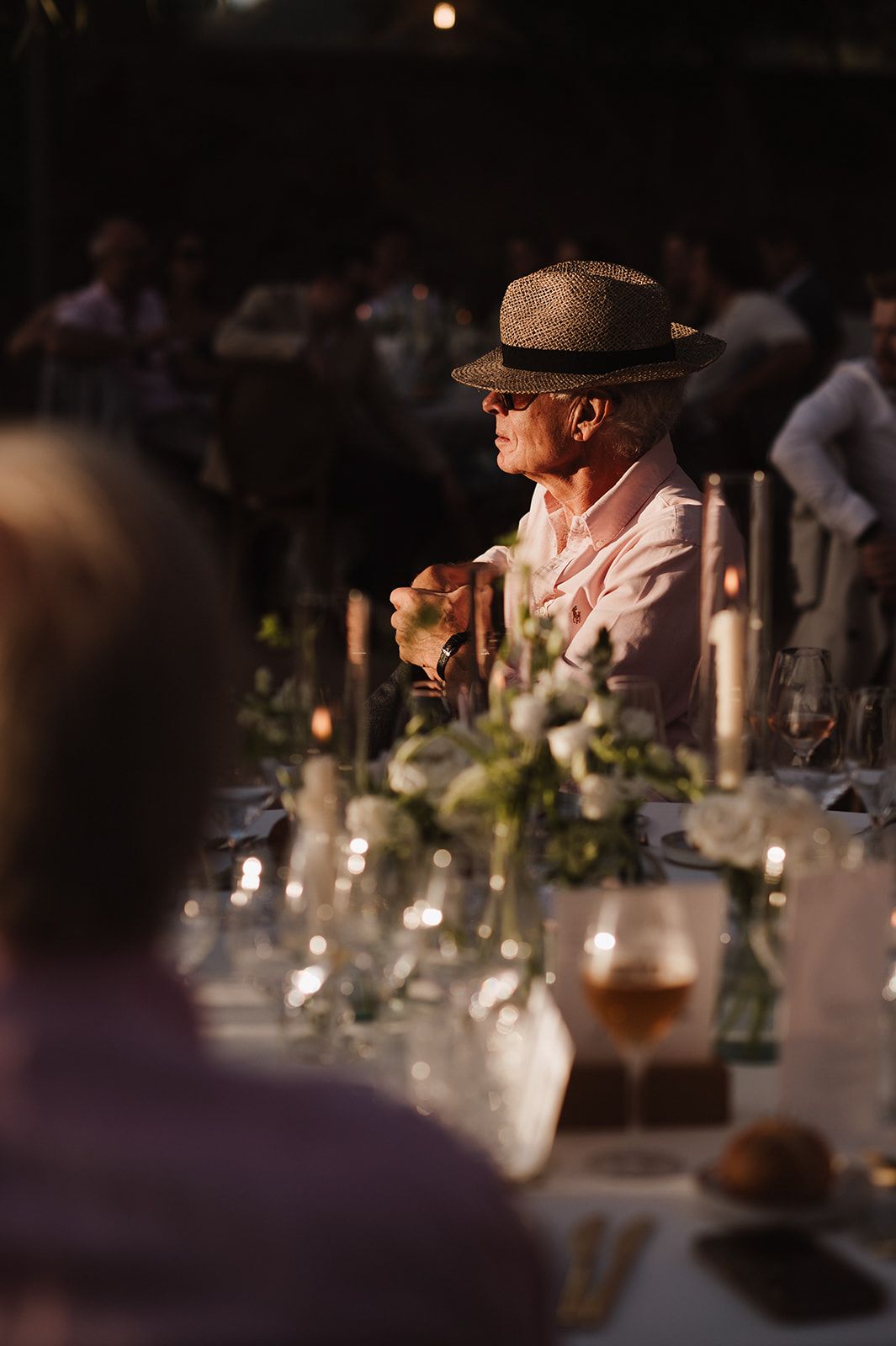 Wedding guest in panama hat seated at candlelit outdoor reception dinner.