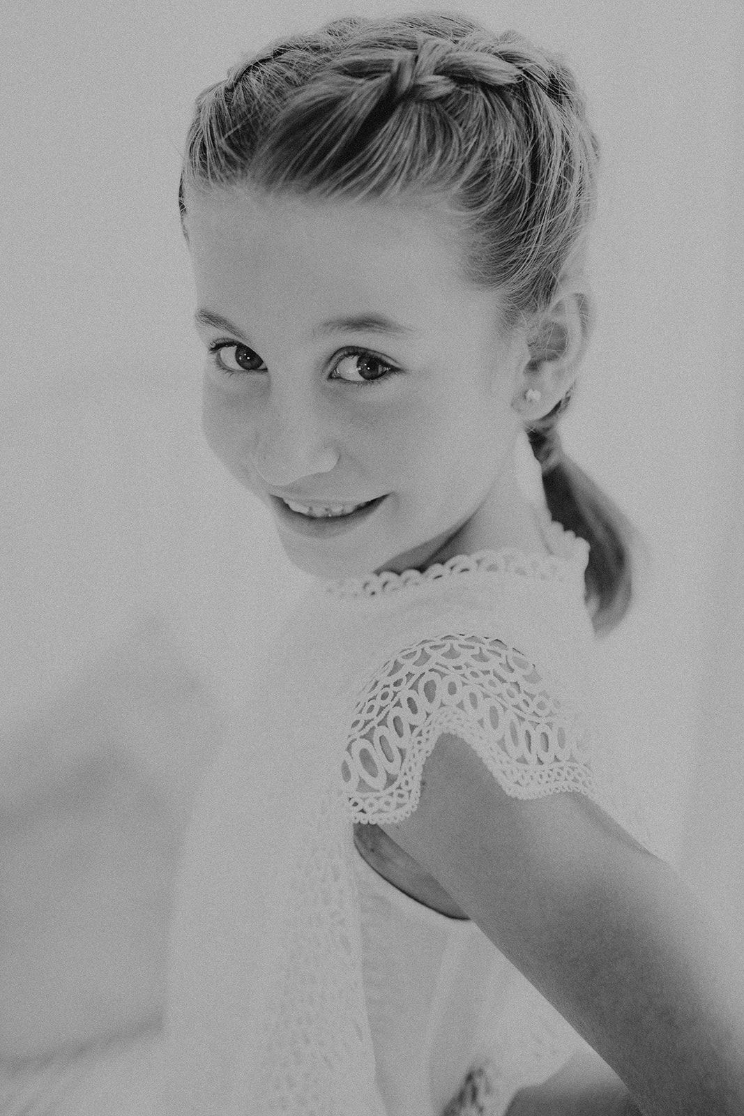 Black and white portrait of a young girl with braided updo hairstyle, wearing a white lace dress, smiling softly.
