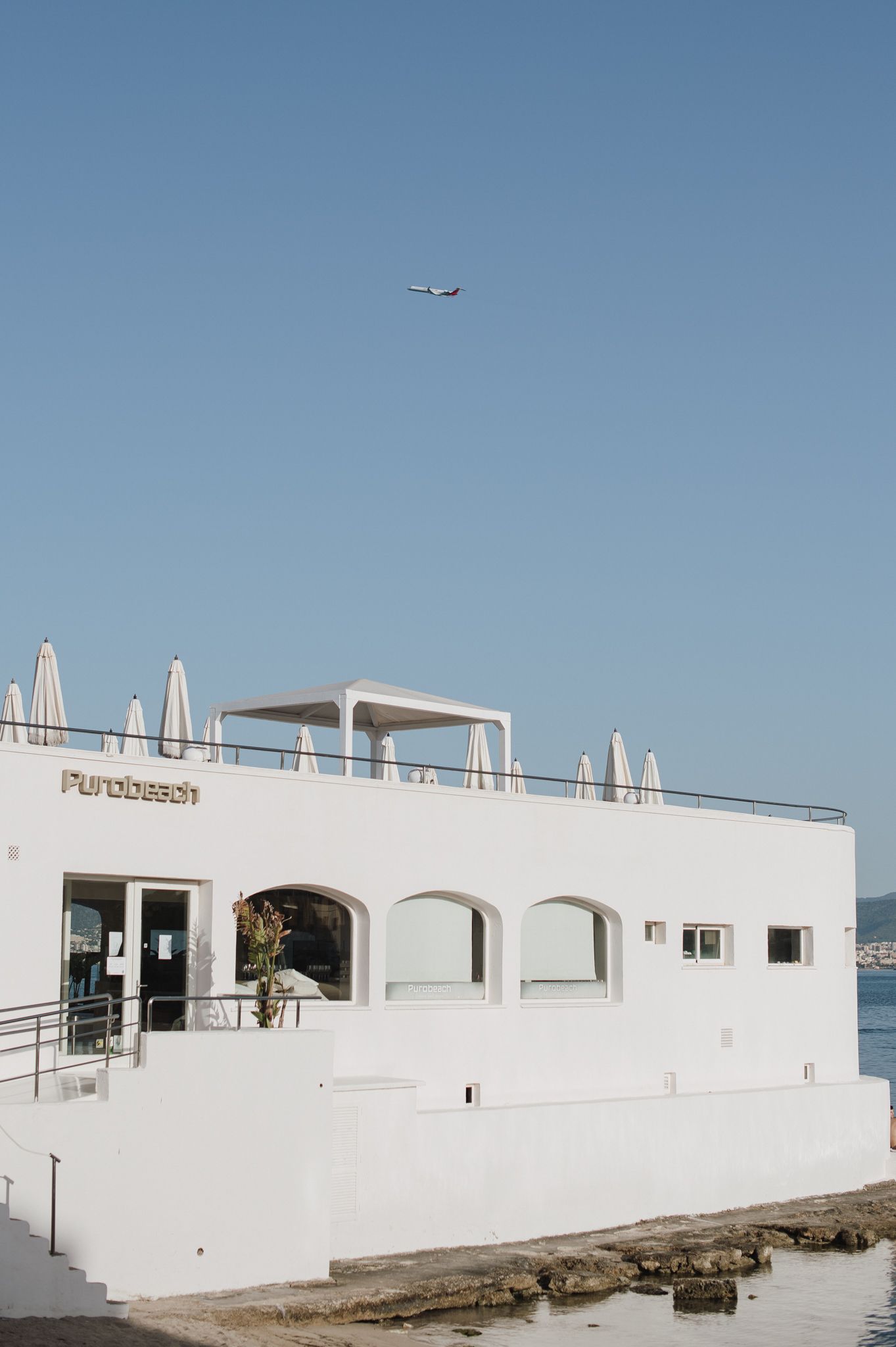 White beachside Purobeach club building with arched windows, rooftop terrace, and airplane in clear blue sky above.