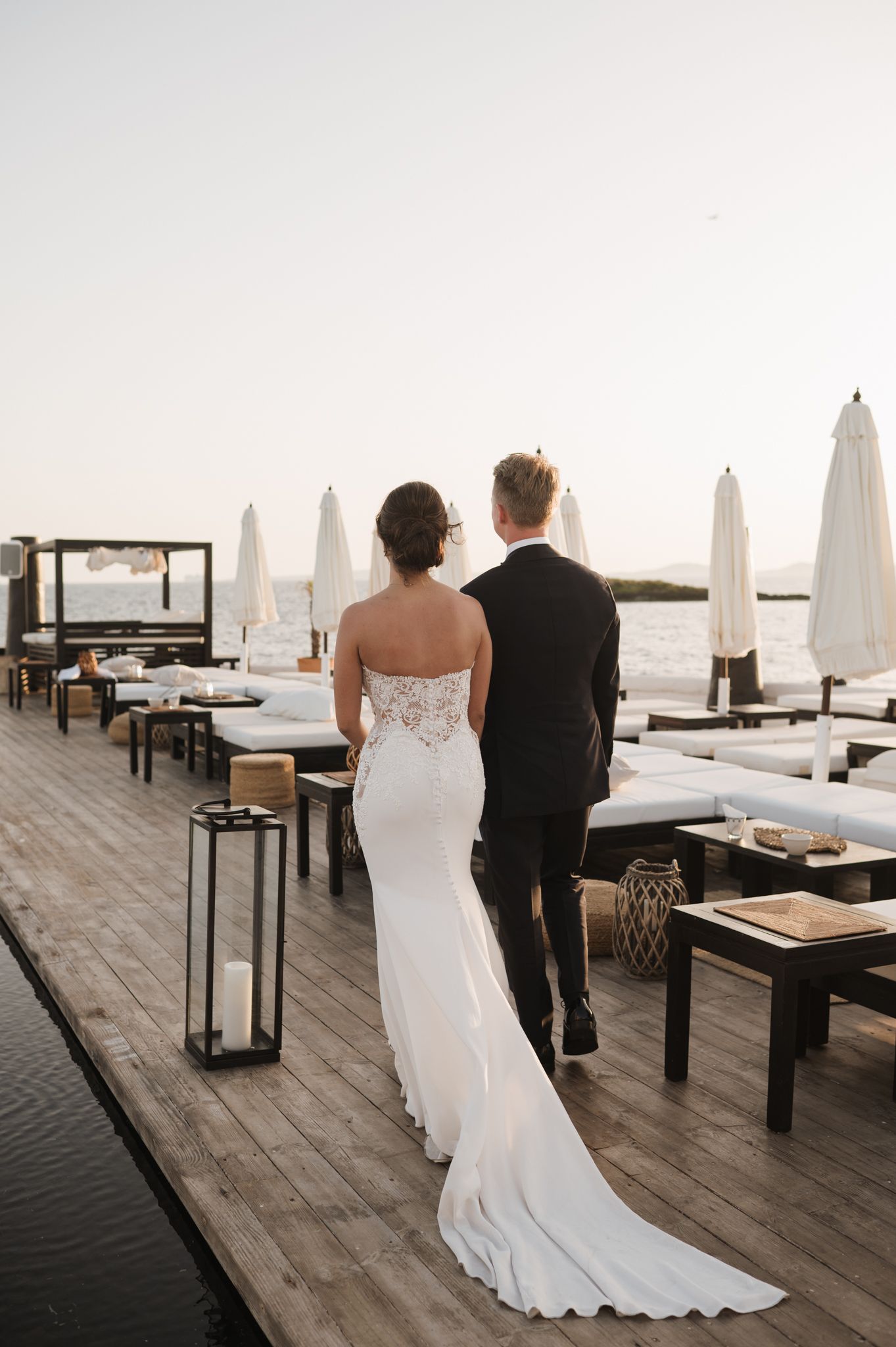 Bride and groom walk hand-in-hand on a waterfront deck at sunset, surrounded by elegant white umbrellas.