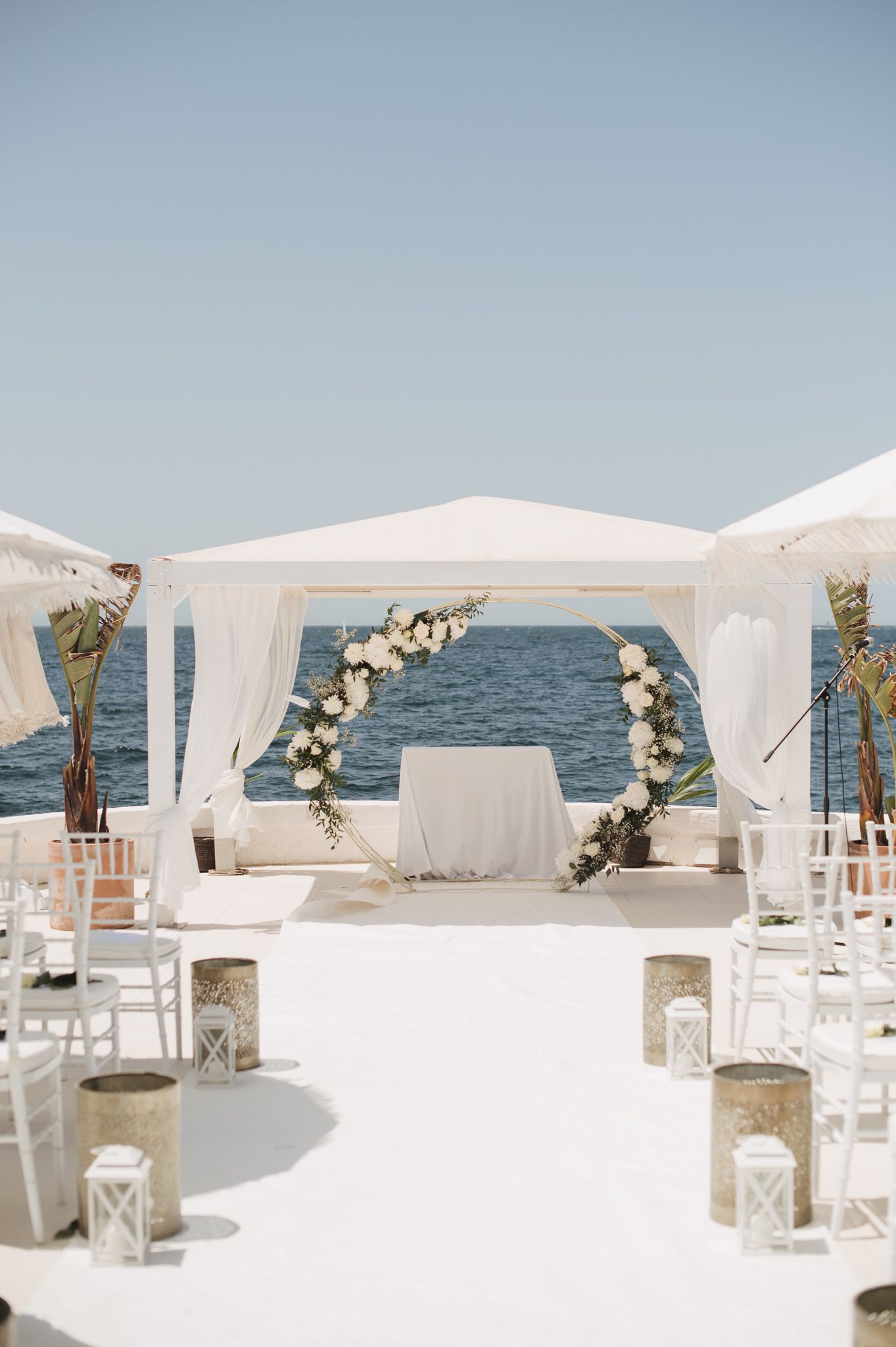 Elegant beachfront wedding ceremony setup with white floral arch, chiavari chairs, and ocean backdrop.