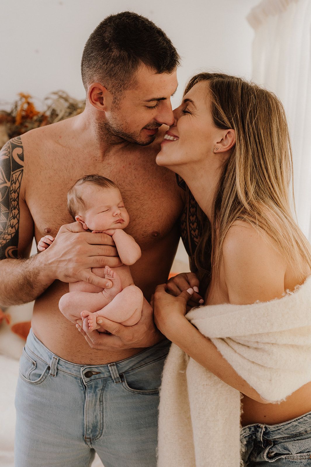 Tattooed shirtless man and blonde woman share a kiss while tenderly holding their sleeping newborn baby.