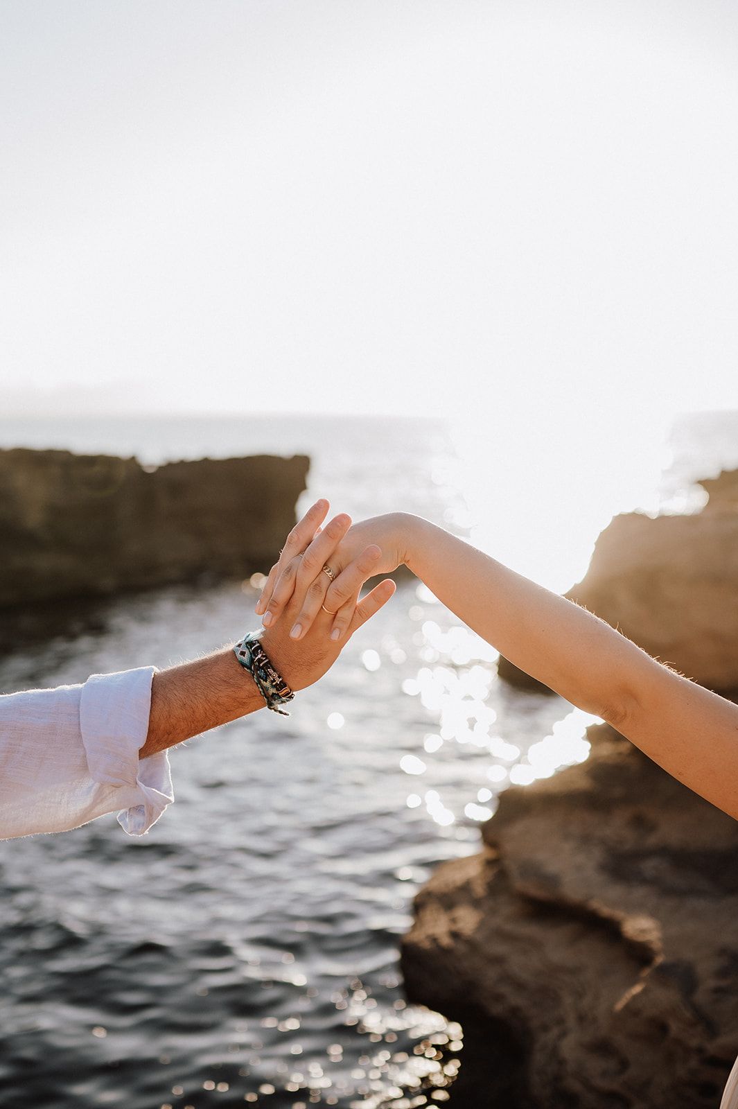 Close-up of couple holding hands with engagement ring visible against sparkling ocean sunset.