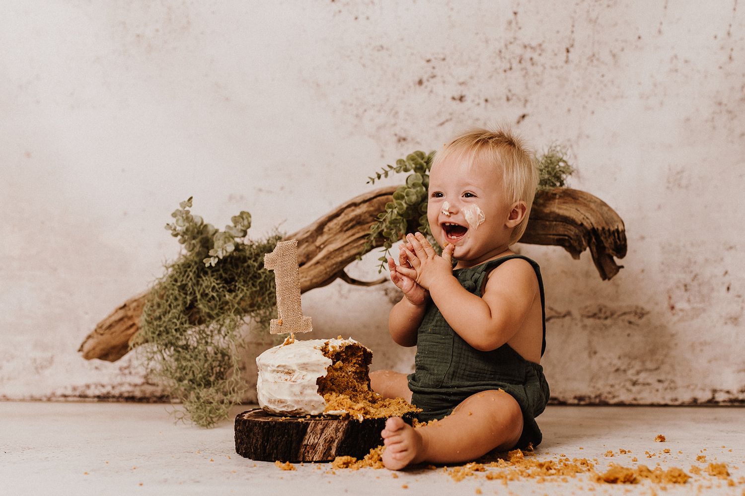 Happy baby in green romper laughing during cake smash photo session with rustic wood backdrop.