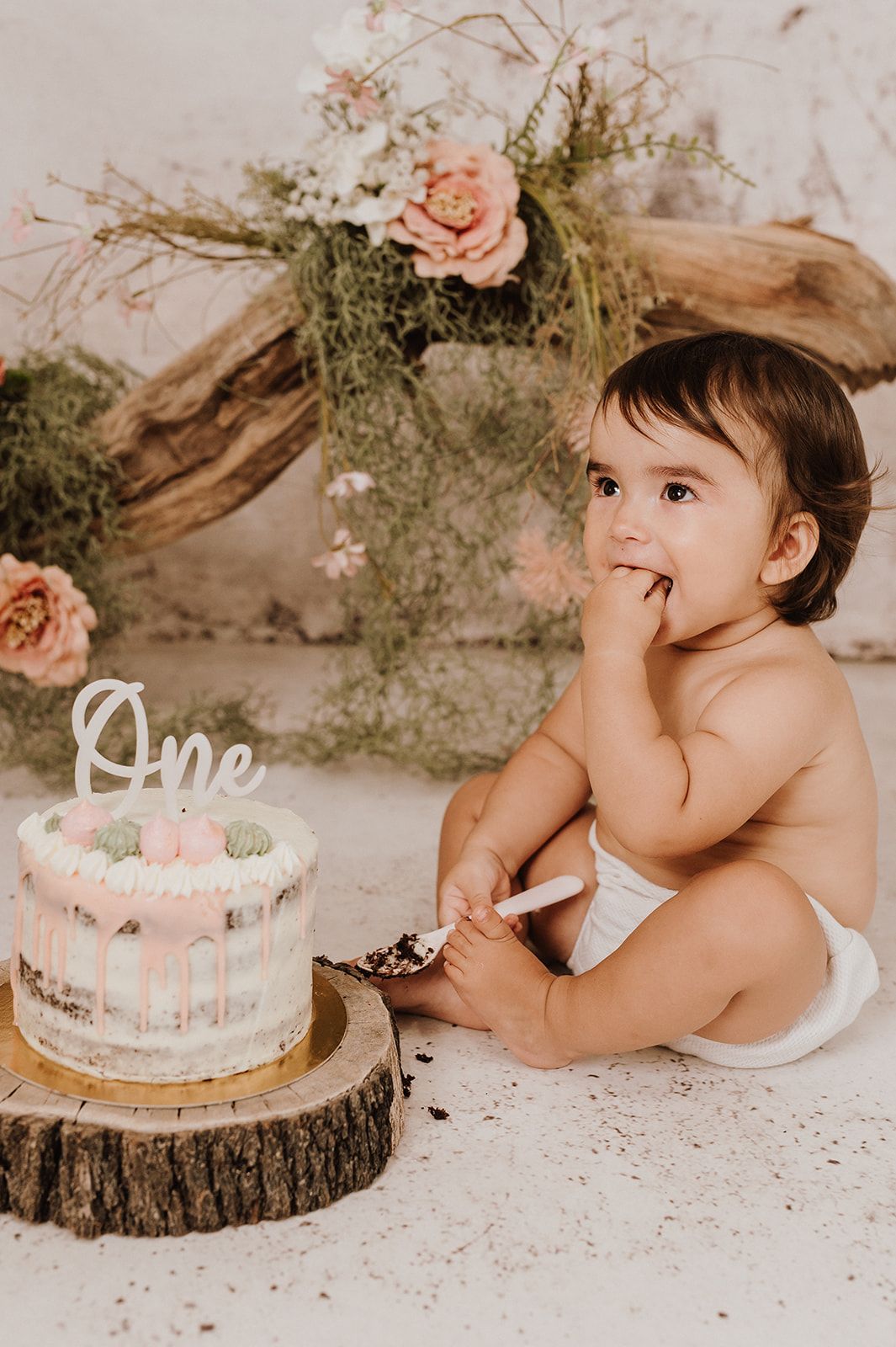 Adorable baby in diaper enjoying first birthday cake smash with pink floral backdrop and wood slice.