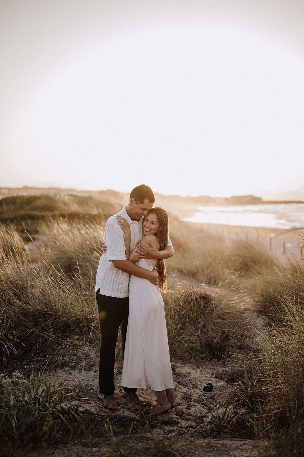 Couple embracing tenderly in coastal grass dunes during a warm golden sunset.
