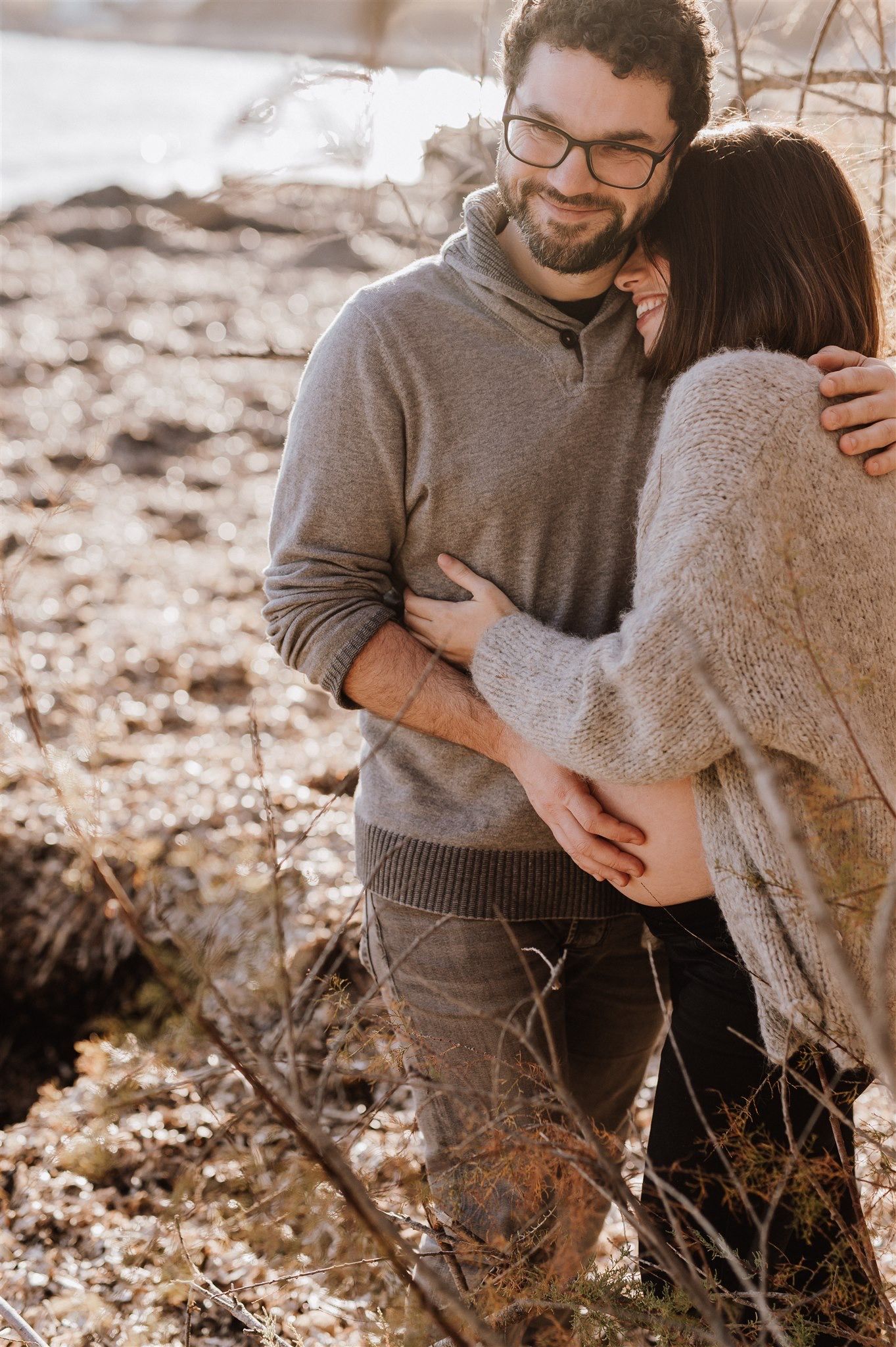 A smiling couple embraces outdoors in autumn, man in gray sweater with glasses hugs woman in cream knit sweater.