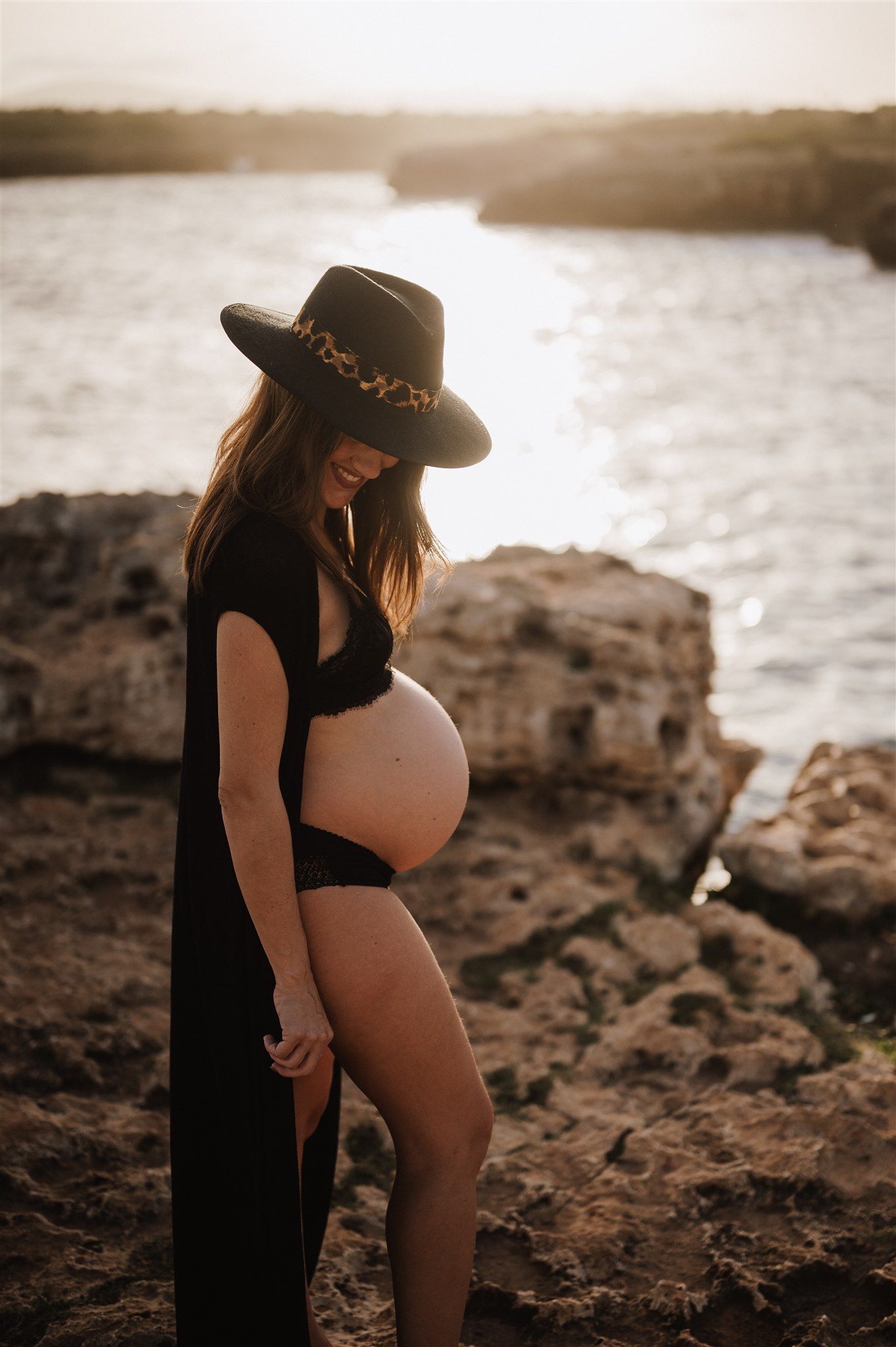 Pregnant woman in black dress and wide-brim hat gazes at belly during golden hour maternity shoot by rocky coastline.