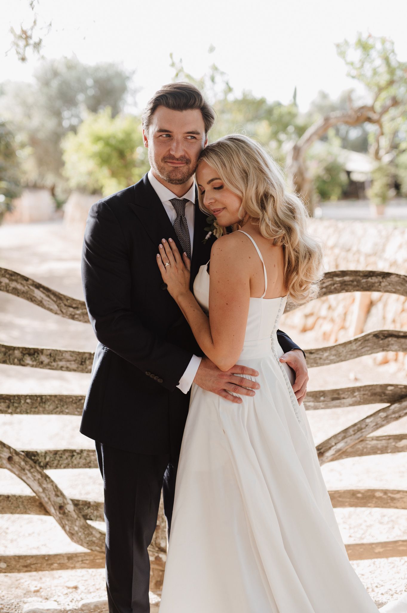 Bride leaning against groom in black suit near a rustic stone fence at their wedding.