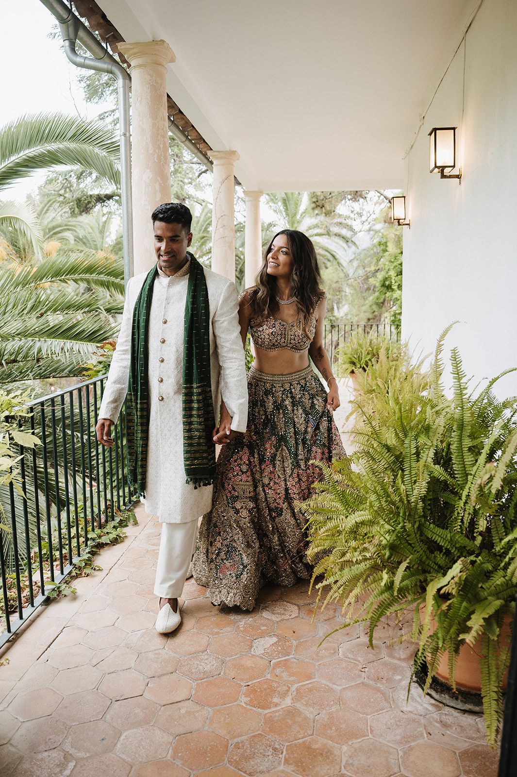 Couple in traditional Indian attire walking on a terrace, man in white kurta with green dupatta, woman in floral lehenga.