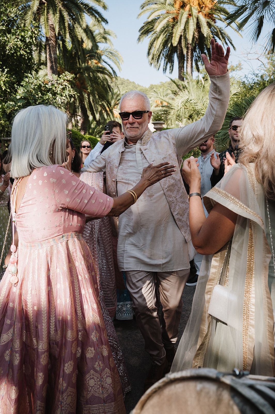 Elderly man dancing enthusiastically surrounded by guests in Indian wedding attire outdoors.