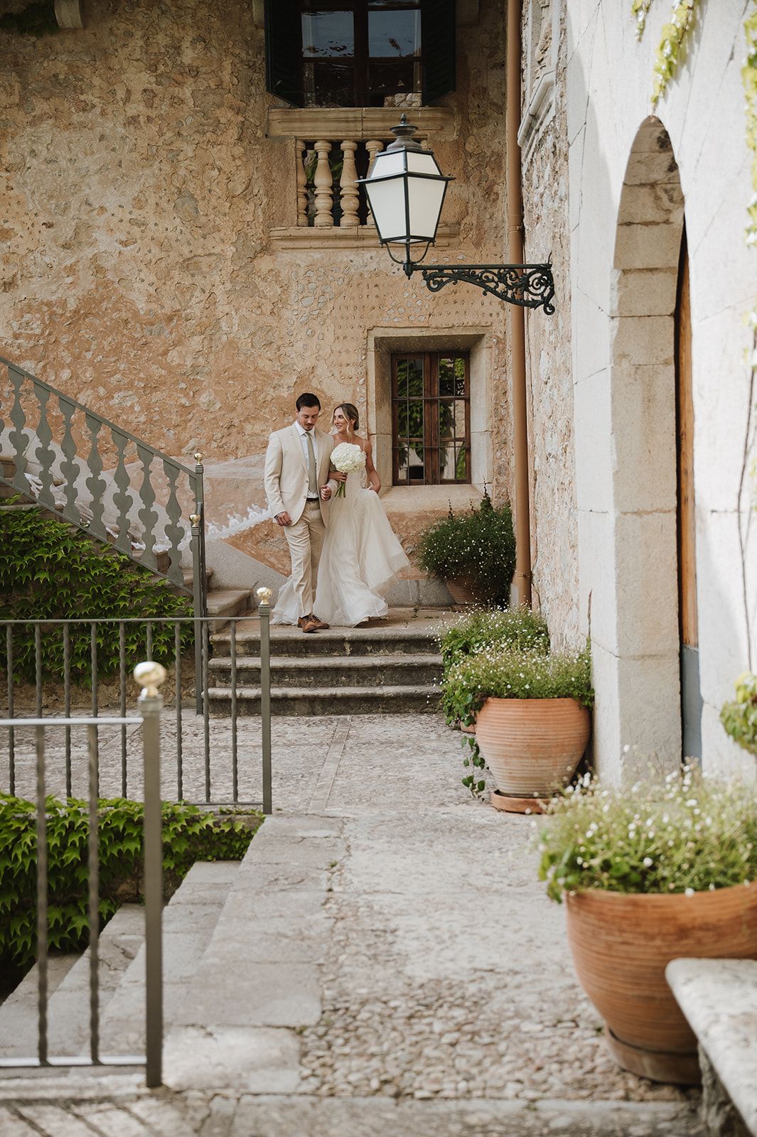 Bride and groom walk together through a rustic stone courtyard with terracotta pots and lush greenery.
