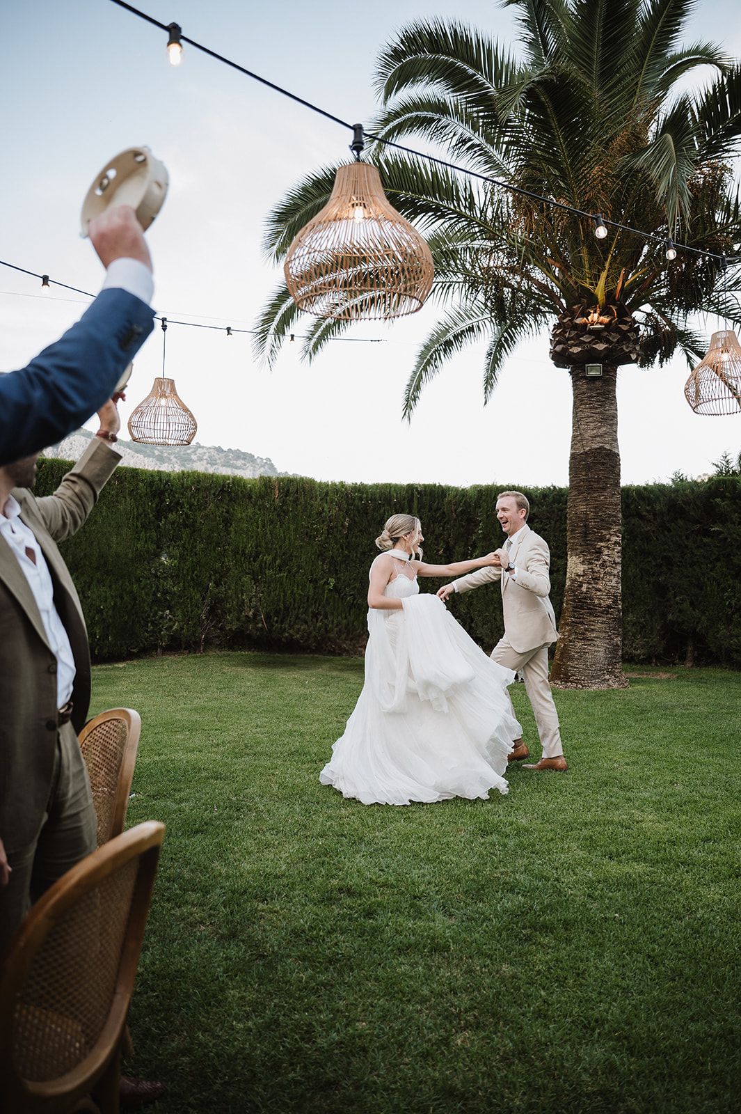 Bride and groom dancing on lush green lawn under string lights and wicker pendant lamps at outdoor wedding.