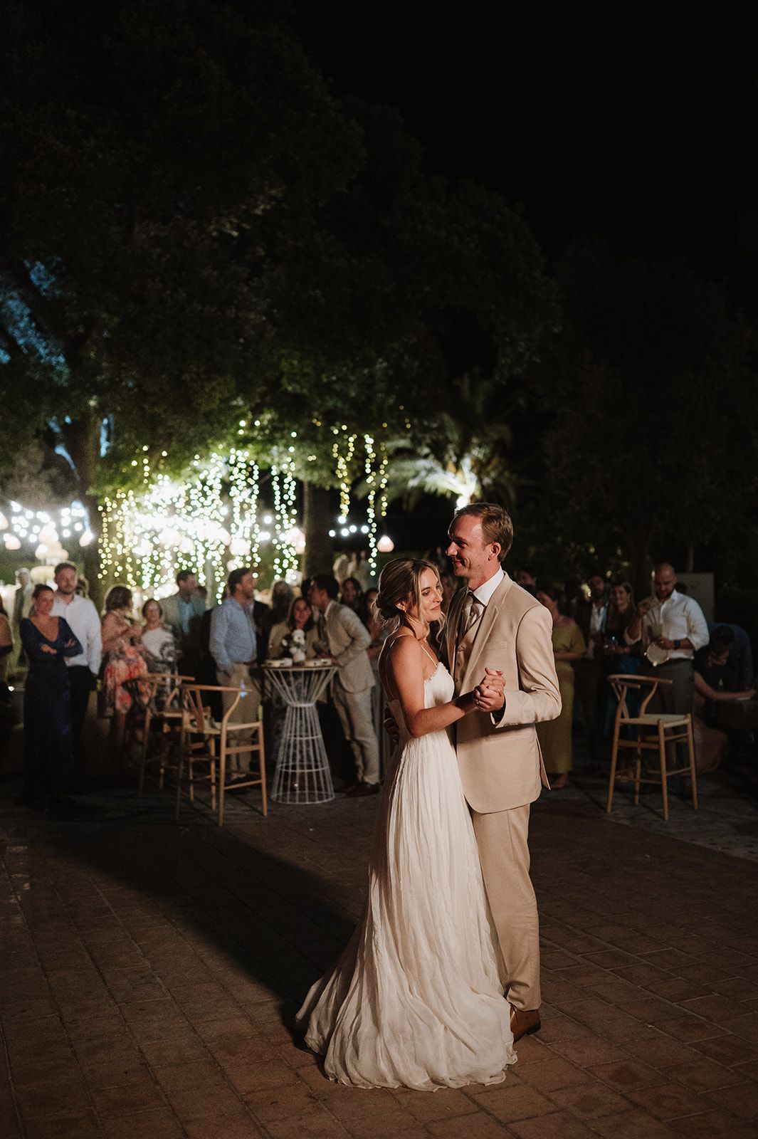 Bride and groom share first dance outdoors at night surrounded by guests and cascading fairy lights.