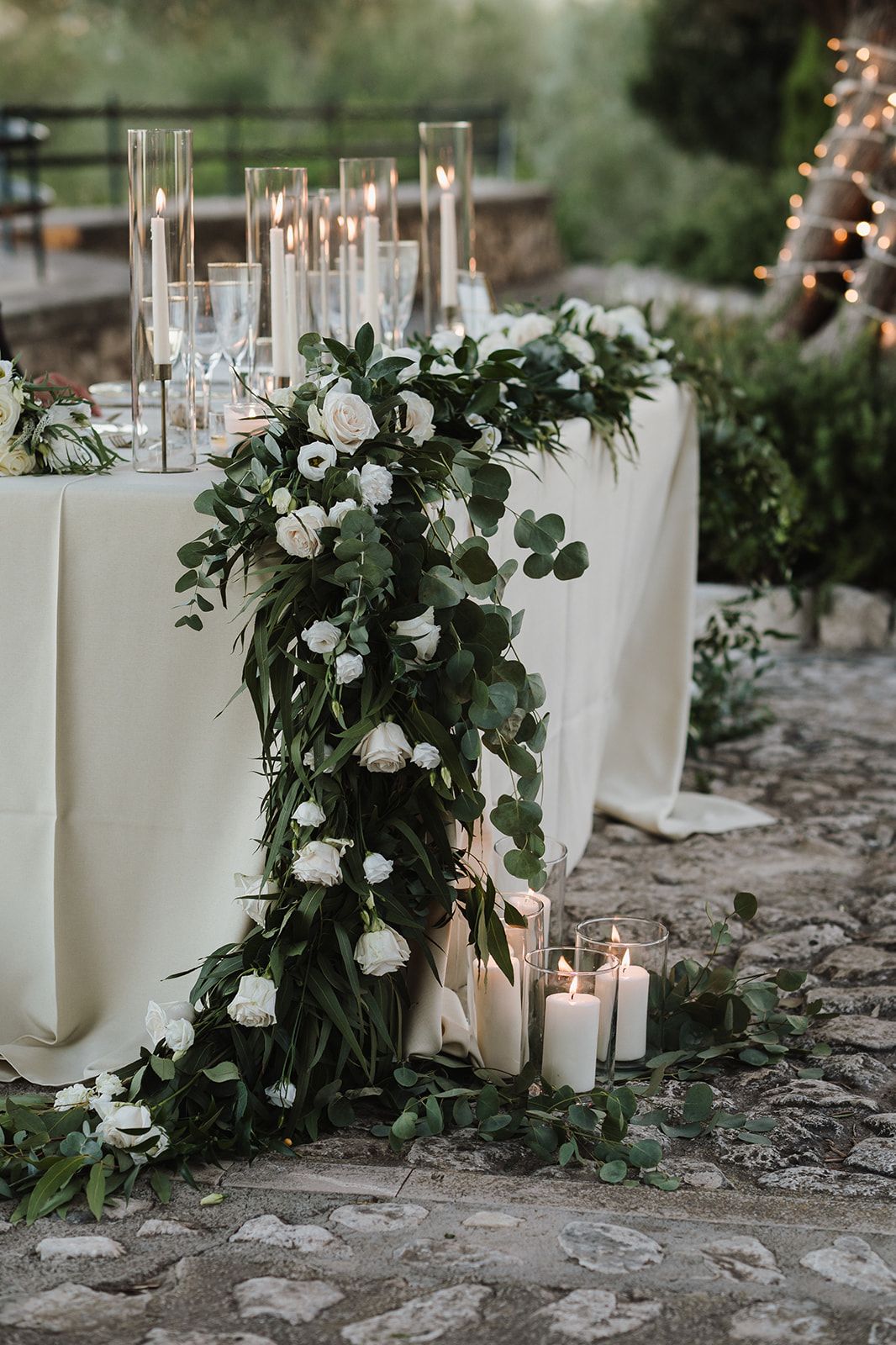 Elegant wedding sweetheart table adorned with white roses, eucalyptus garland, and pillar candles on stone floor.