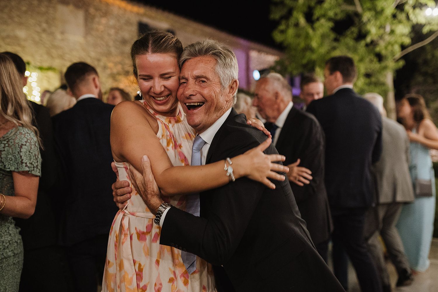 Joyful wedding guests embrace and laugh together during lively outdoor evening wedding reception celebration.