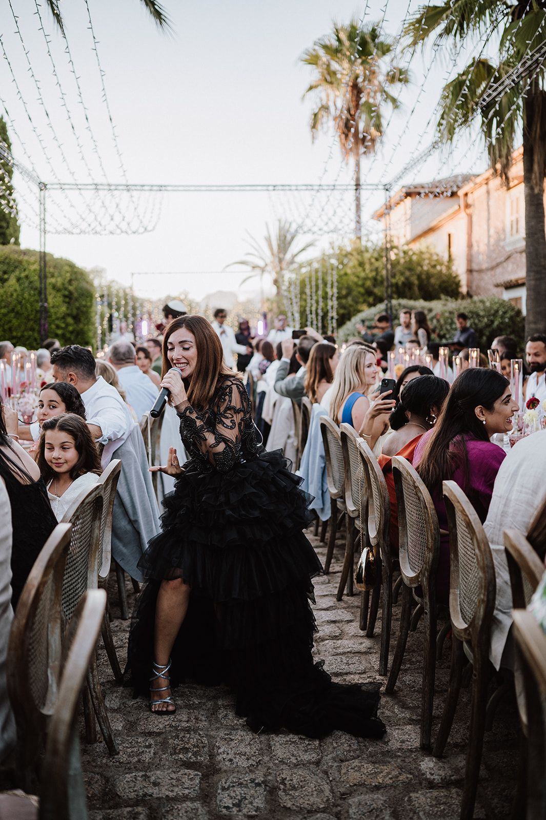Wedding guest in dramatic black ruffled gown walks through outdoor reception venue lined with string lights.