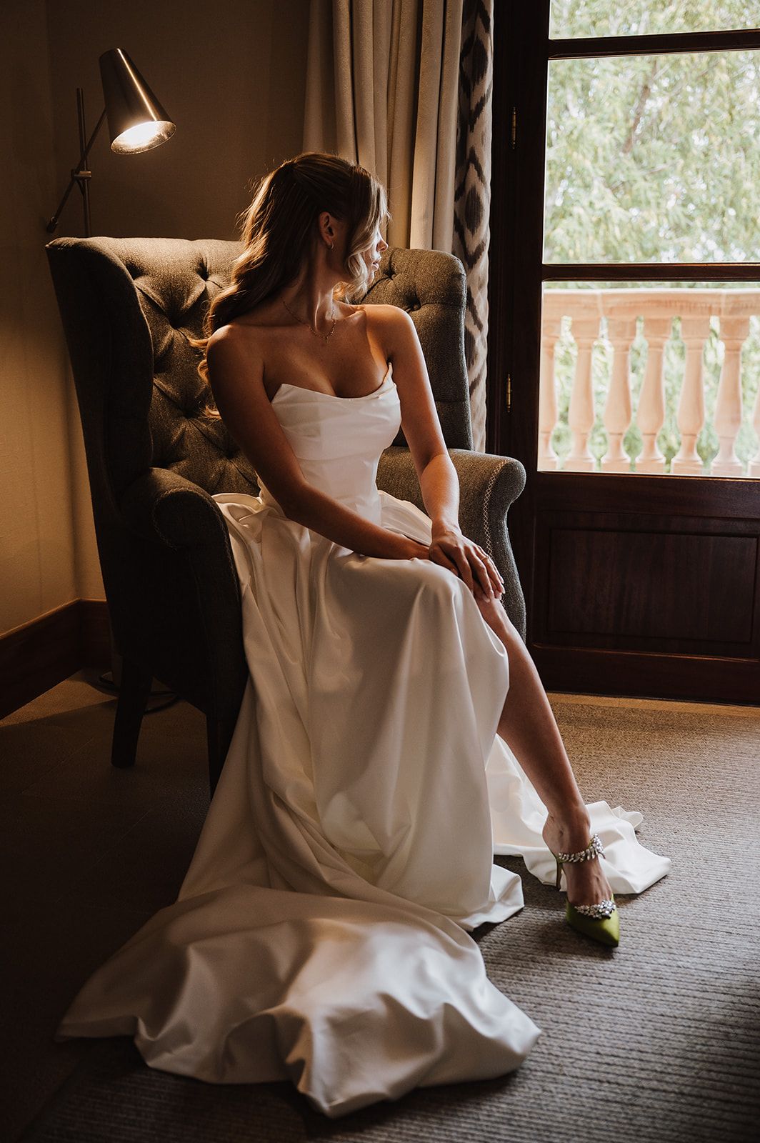 Bride in strapless white wedding gown seated in tufted armchair by window in warm light.
