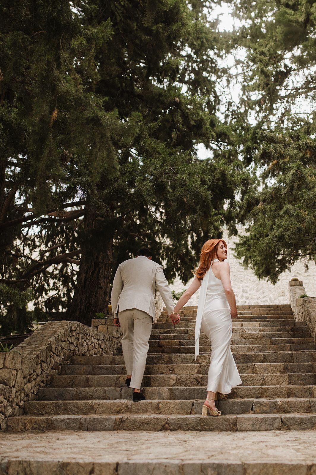 Bride and groom holding hands climbing rustic stone steps beneath tall cypress trees at sunset.