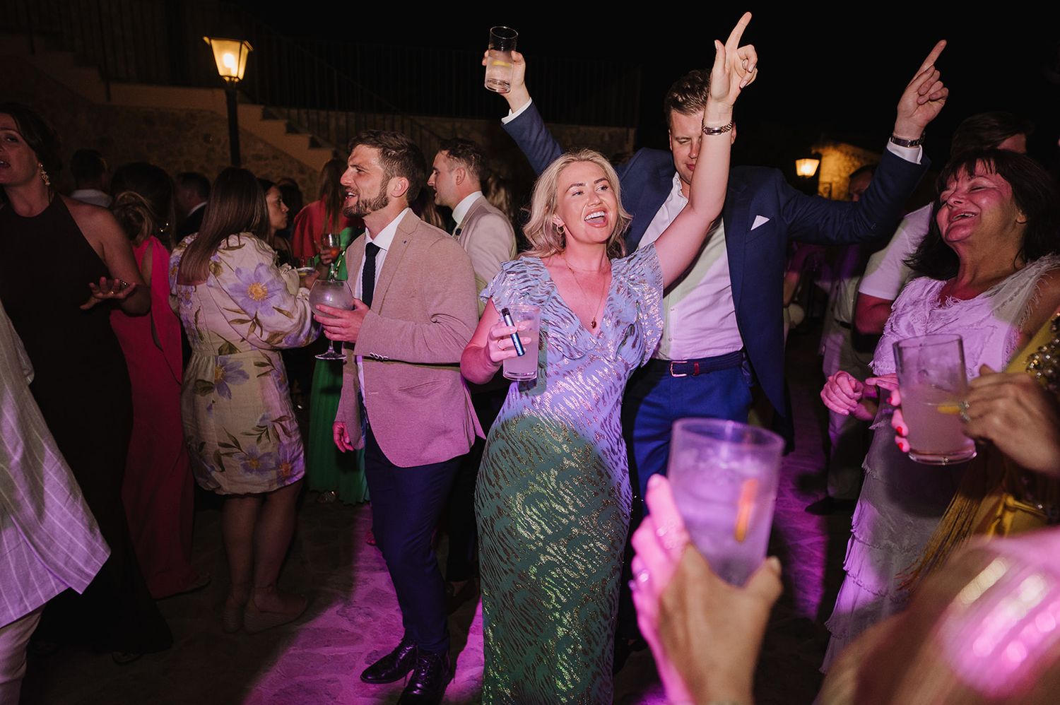 Wedding guests dancing joyfully under pink lights at an outdoor evening wedding reception party.