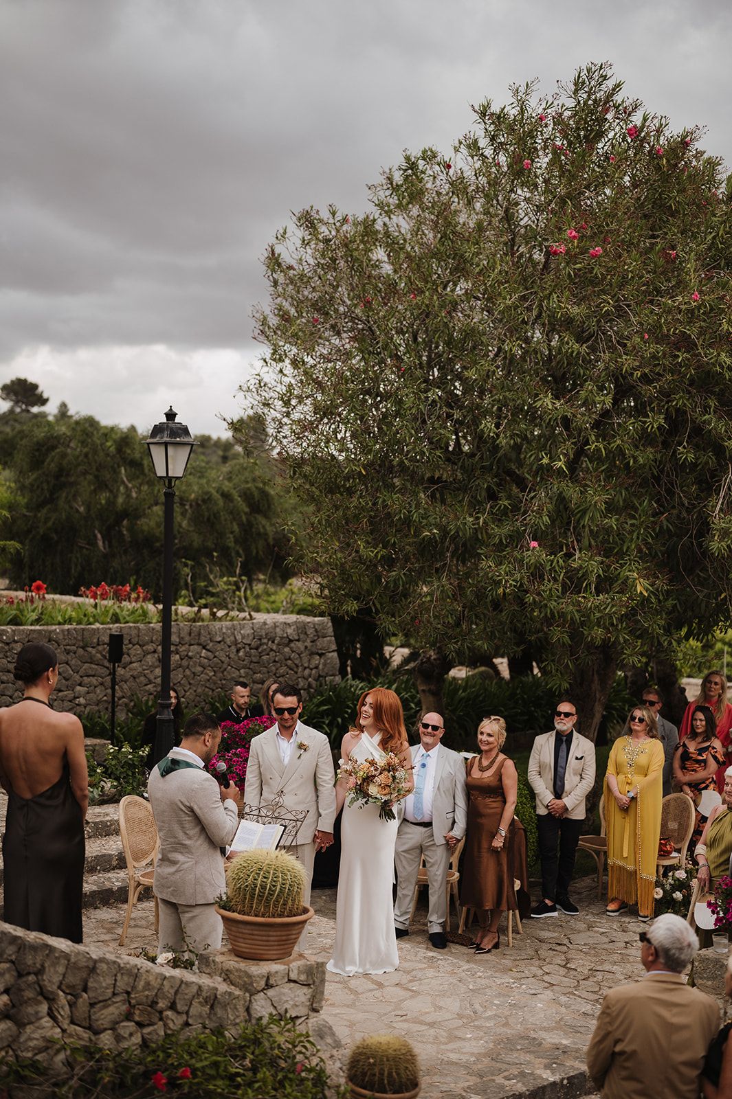 Couple exchanges vows at an outdoor wedding ceremony with lush greenery and stone walls in background.