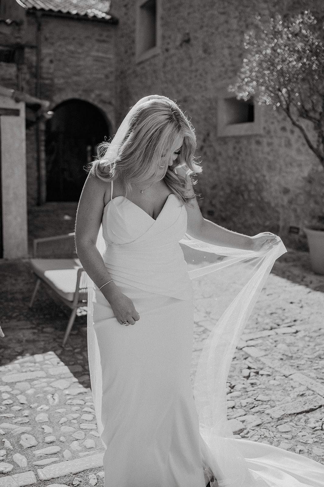 A bride in a flowing white wedding gown holds her veil outdoors at a rustic stone venue in black and white photo.