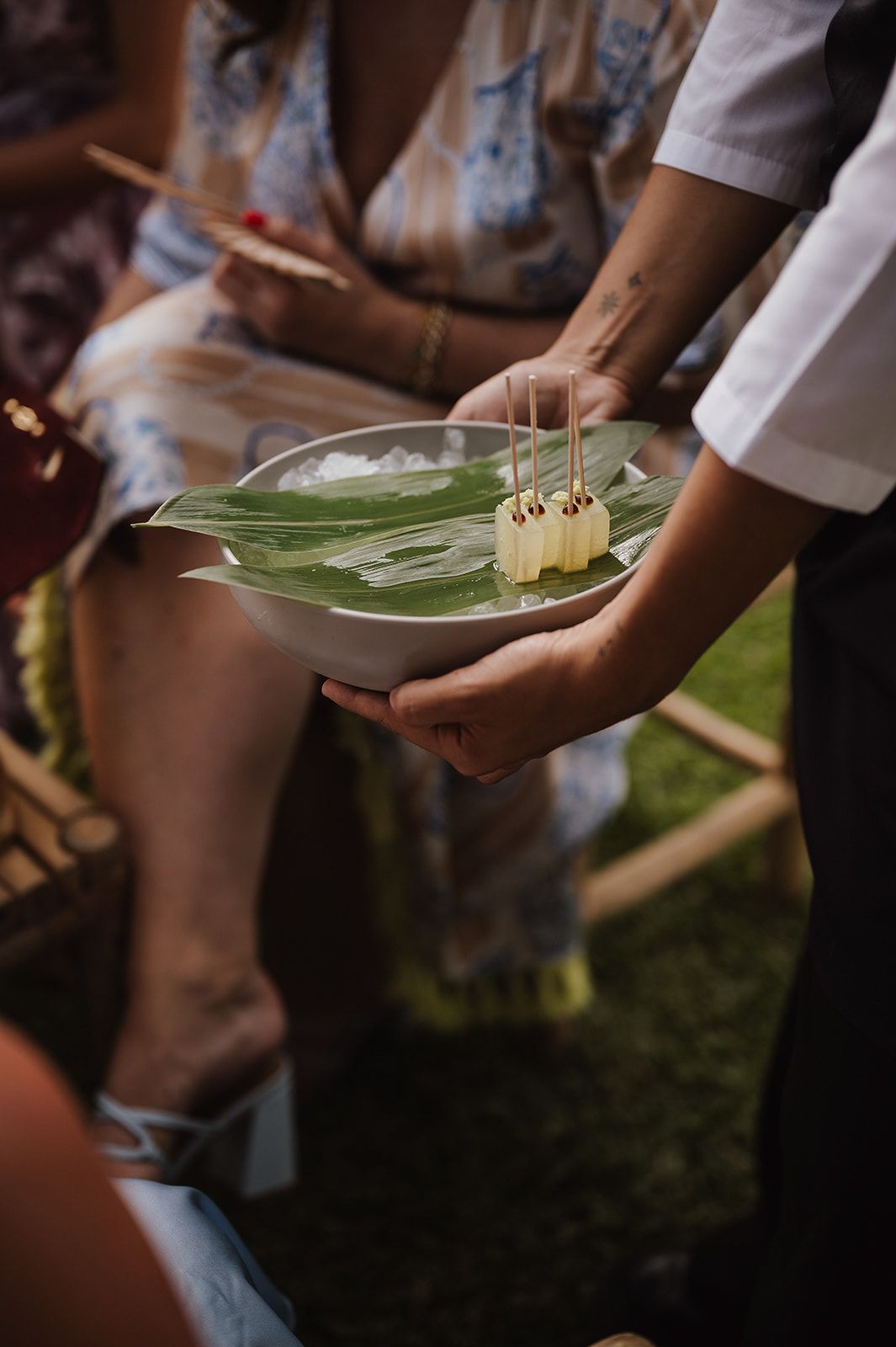 A server presents elegant appetizers on a banana leaf-lined tray at an outdoor wedding reception.
