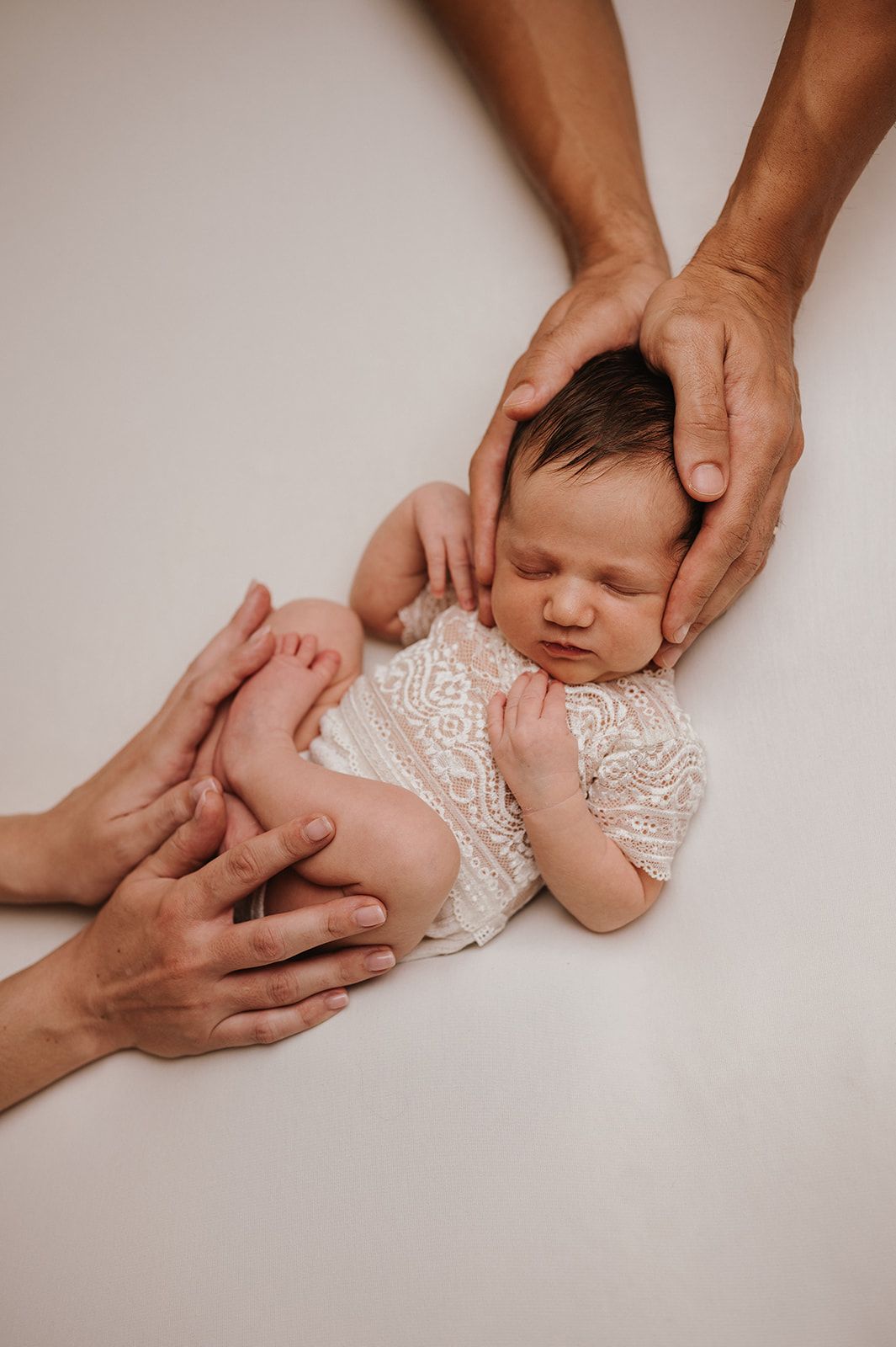 Newborn baby in white lace outfit sleeping peacefully while parents gently cradle with loving hands on white background.