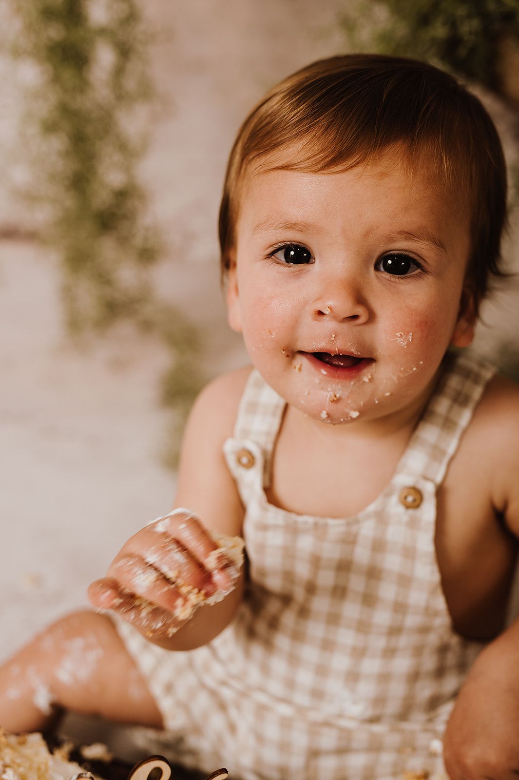 Adorable baby in gingham overalls with cake smash mess on face, smiling during first birthday photo session.