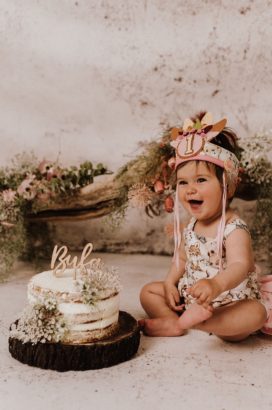 Baby girl in floral romper celebrating first birthday with naked cake topped with Byfa topper and dried flowers.