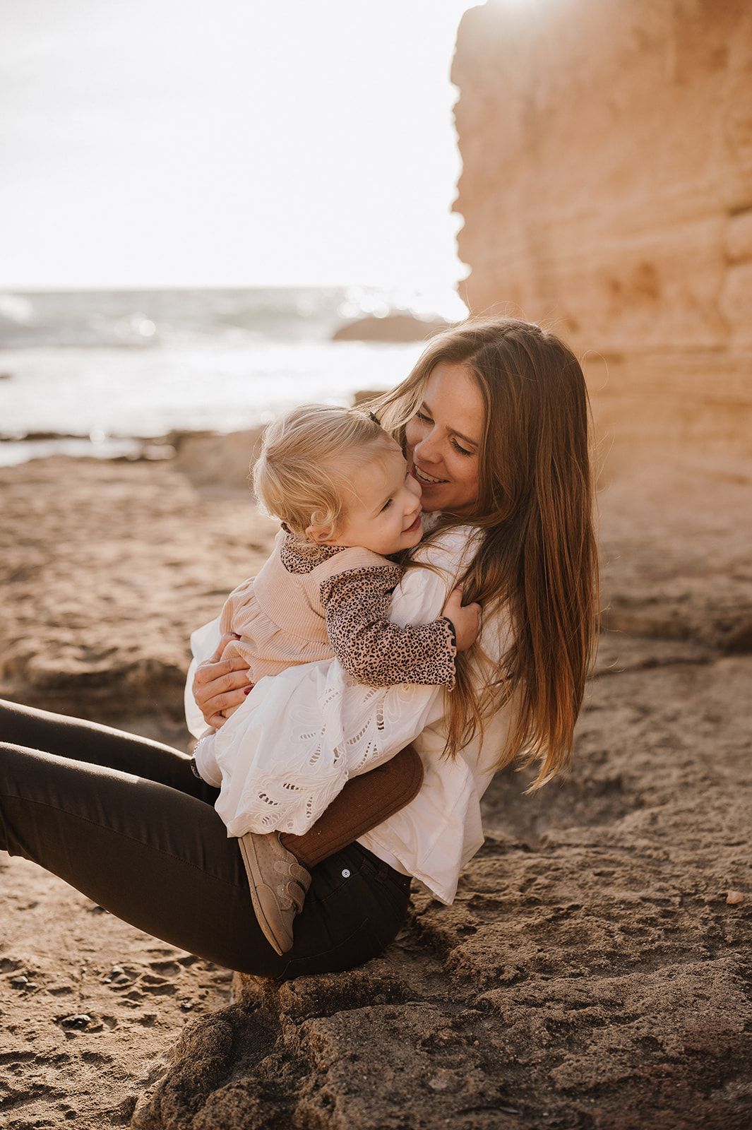A loving mother sits on sandy beach rocks cradling her young daughter in warm golden sunlight.